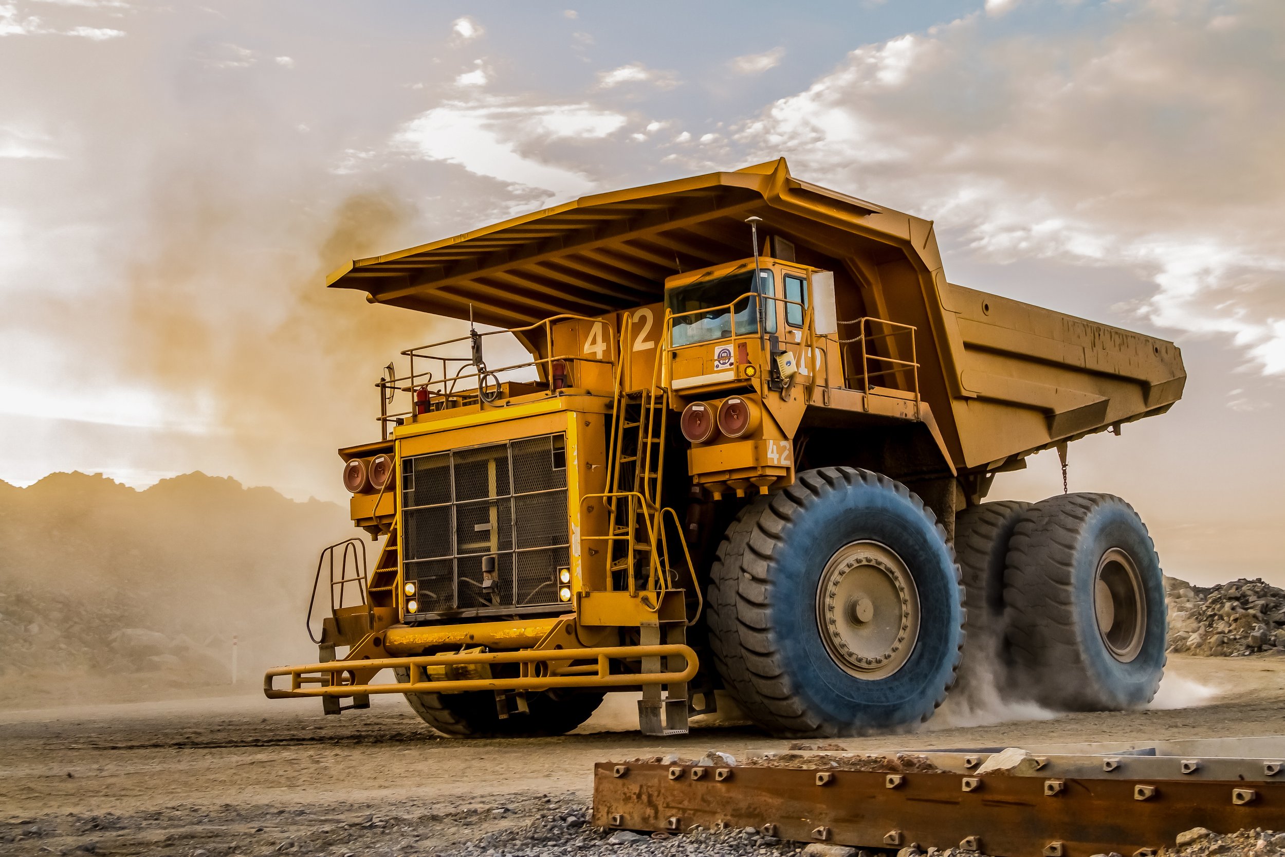 A large yellow dump truck is moving on a dirt road, kicking up dust against a cloudy sky. The truck has massive tires and a wide open cargo bed, with numbers 42 visible on the side.