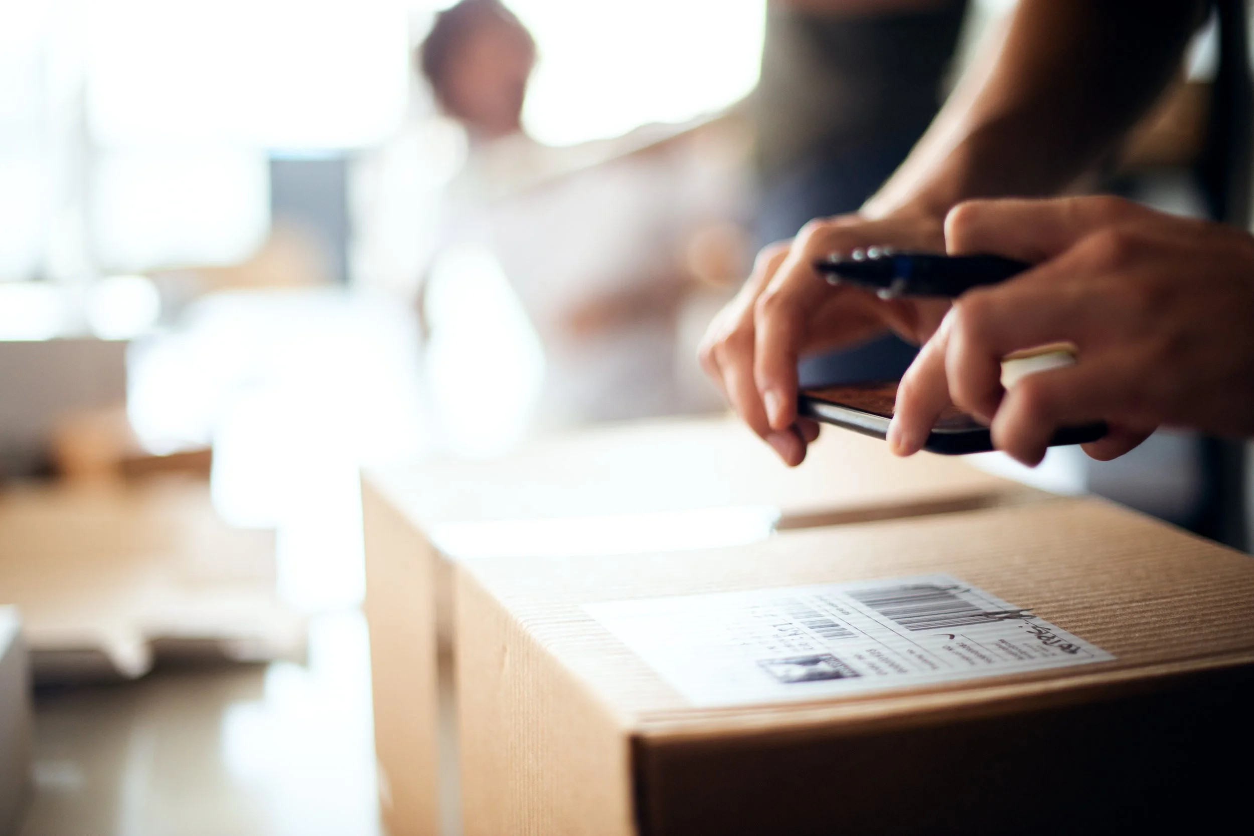 Close-up of hands holding a smartphone over a cardboard box with shipping labels, with a blurred person in the background.