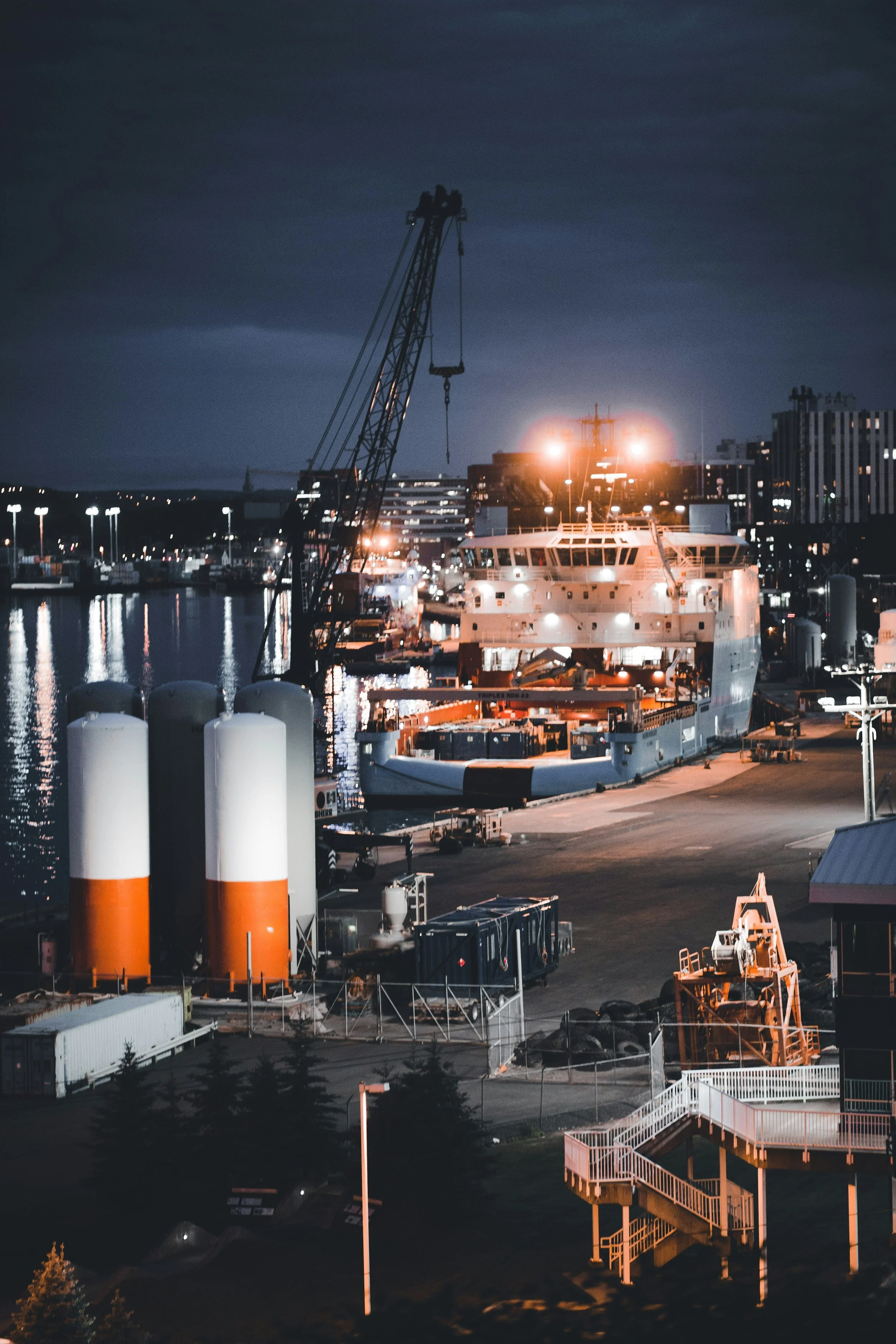 Nighttime view of a marina with a large cruise ship docked, bright lights on the ship and around the area, industrial equipment including large tanks and a crane, buildings in the background, and an emergency staircase illuminated by artificial lighting.