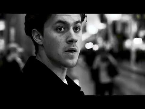 Black and white photo of a young man looking back over his shoulder with a serious expression, standing outdoors in an urban setting.