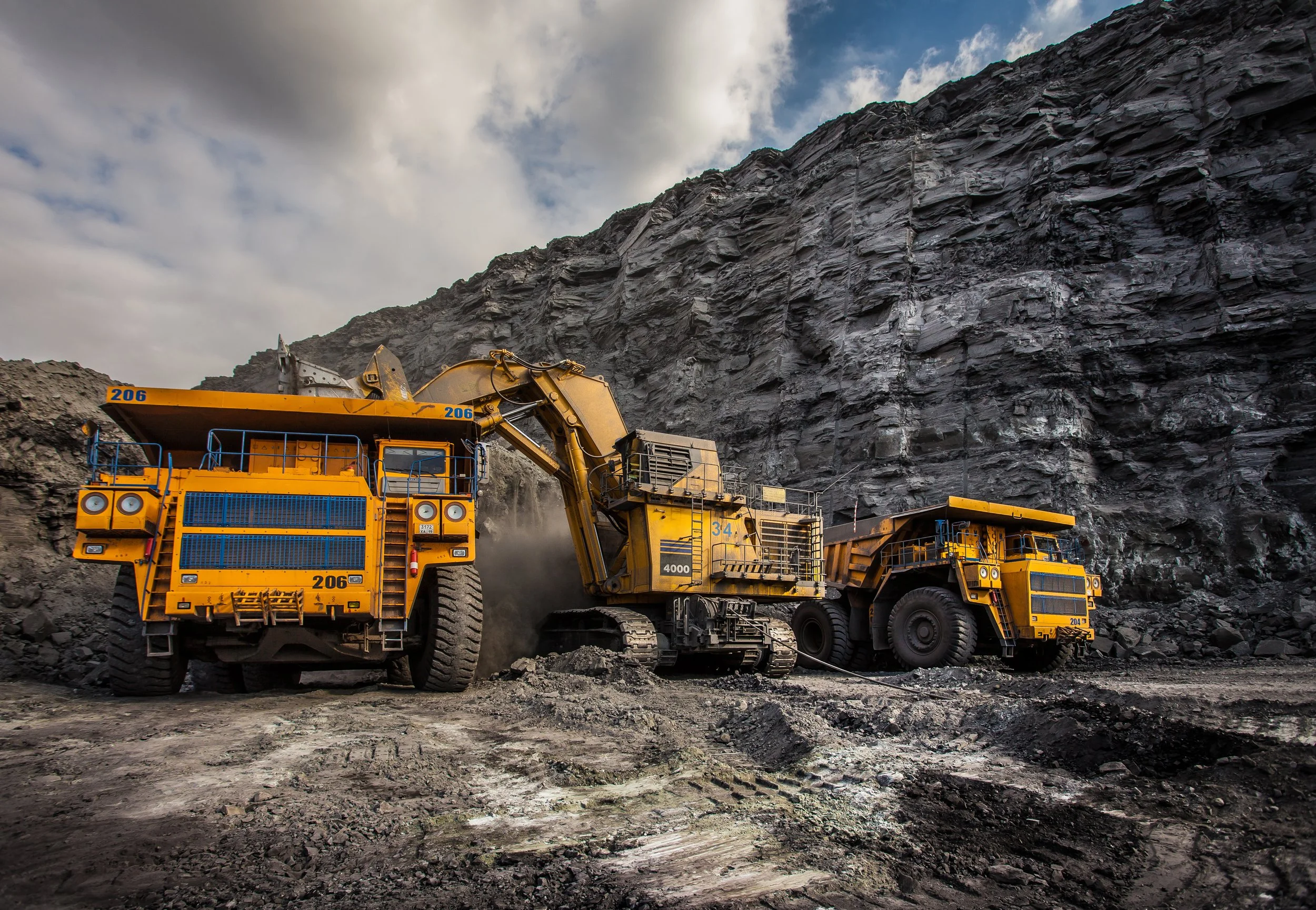 Construction site with large yellow mining trucks operating near a rocky cliff under cloudy sky.