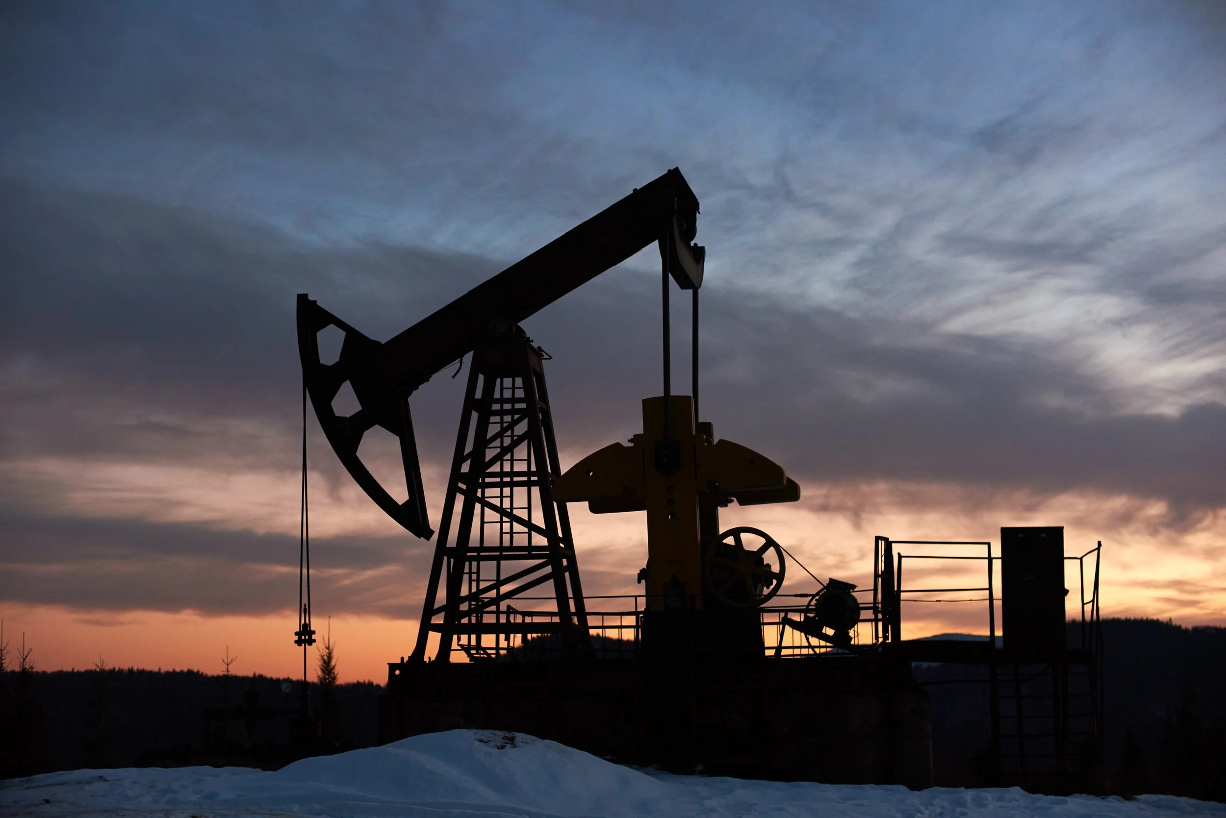 Silhouette of an oil pumpjack against a sunset sky with clouds, with snow on the ground.