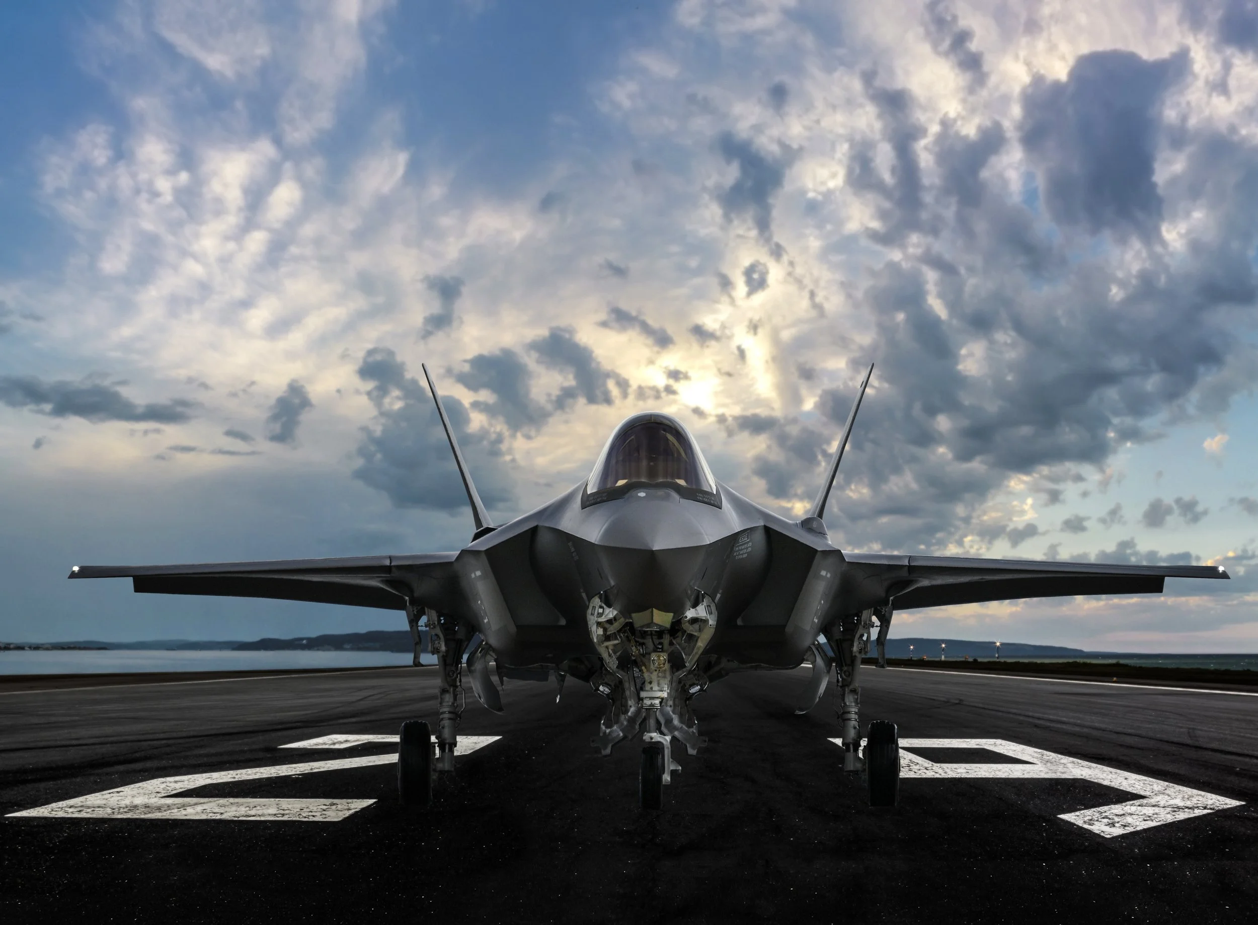 Front view of a fighter jet aircraft on a runway with painted white markings, against a partly cloudy sky at dusk or dawn.