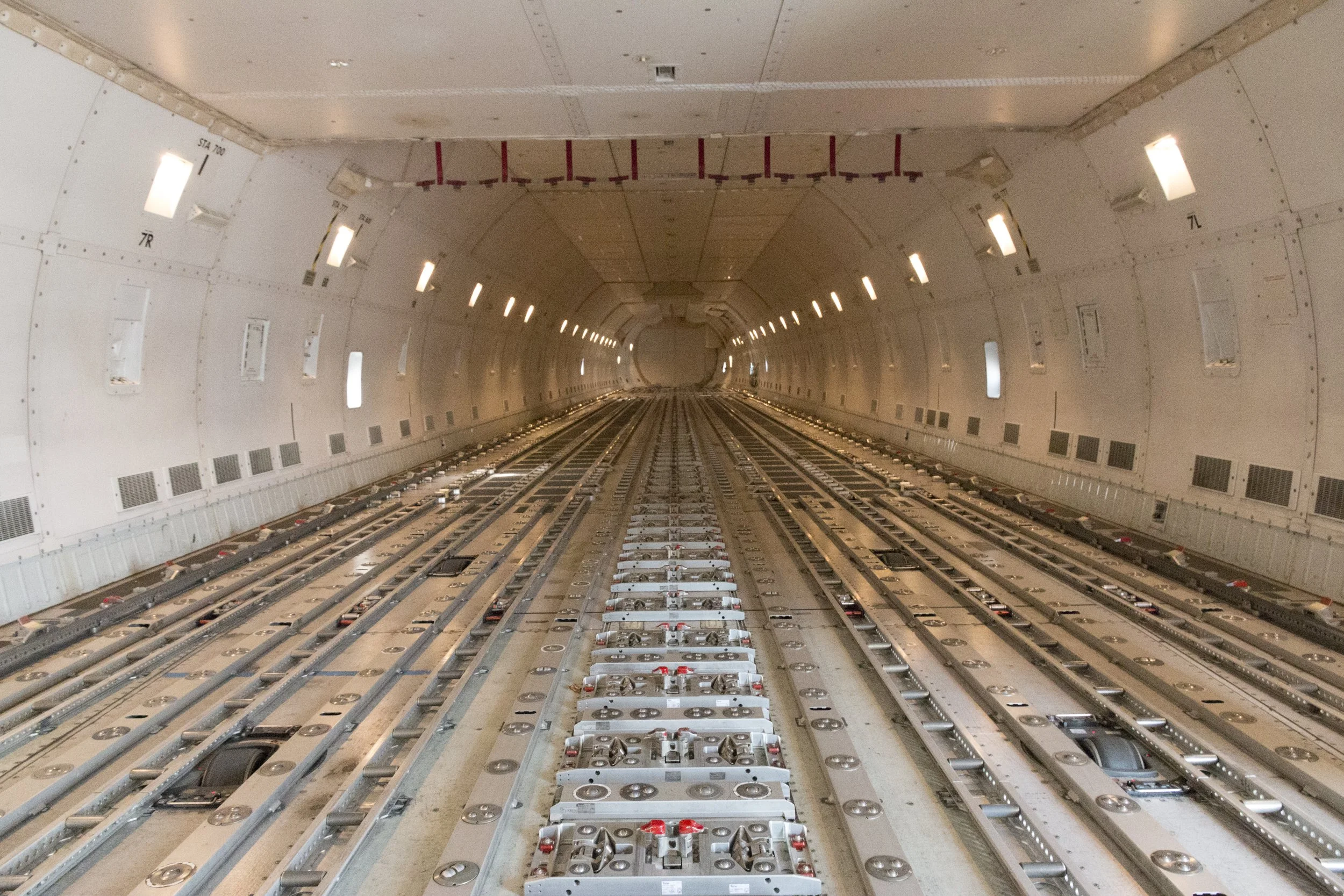 Interior of an aircraft or train tunnel showing exposed structural framework and lighting fixtures along the ceiling and tracks along the floor.
