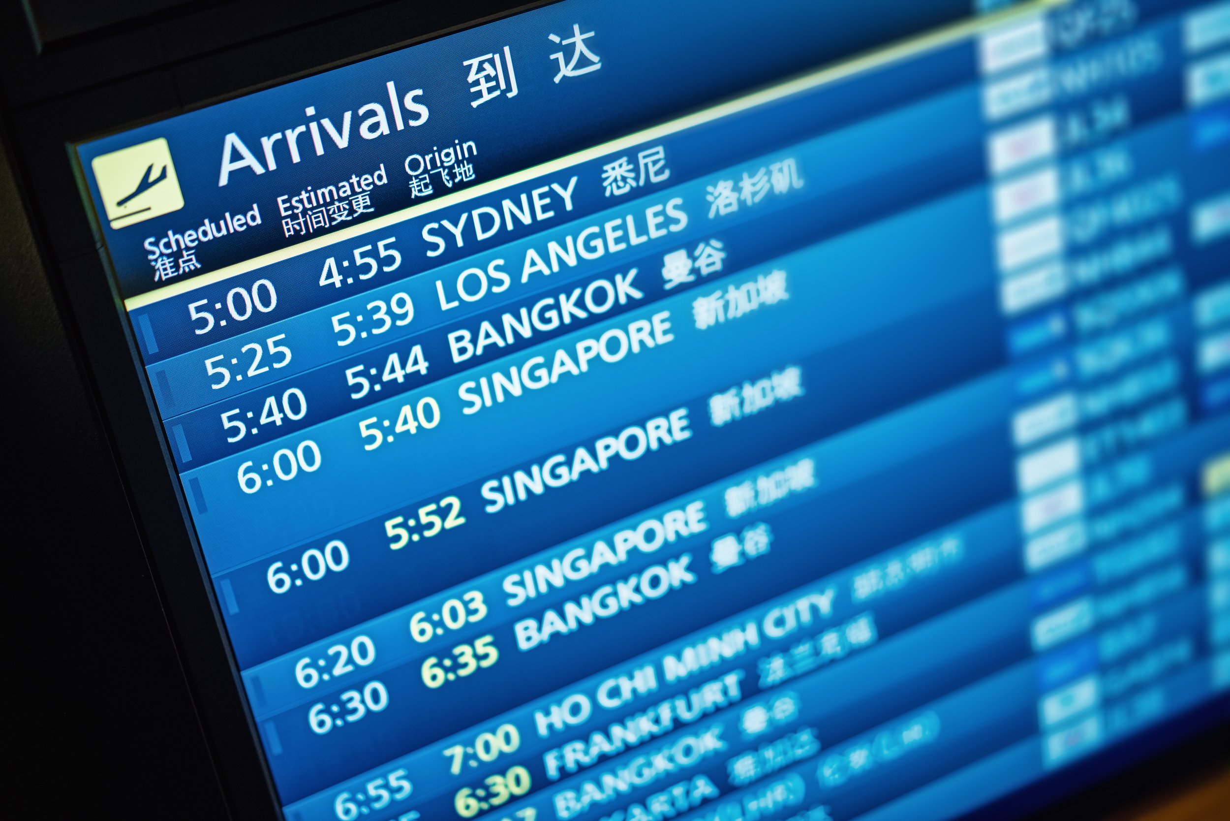 Digital airport arrivals board showing flight times and destinations, including Sydney, Los Angeles, Bangkok, Singapore, and other cities, with some information in Chinese characters.