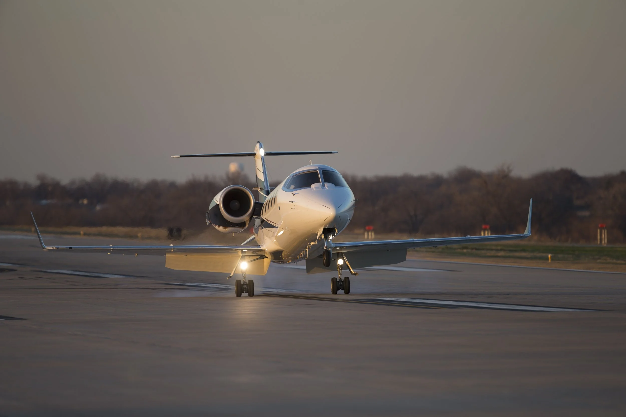 Front view of a sleek white private jet on an airport runway with trees and a cloudy sky in the background during sunset.
