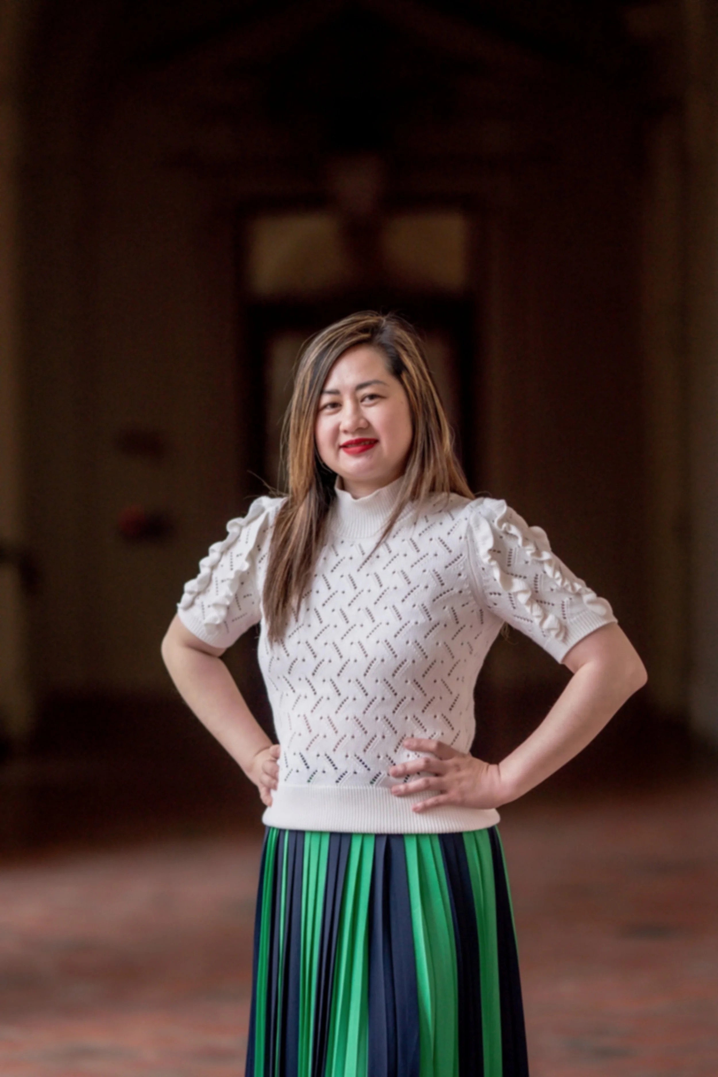 A woman with long brown hair, wearing a white patterned sweater and a green and blue striped skirt, stands with her hands on her hips, smiling in a blurred indoor background.