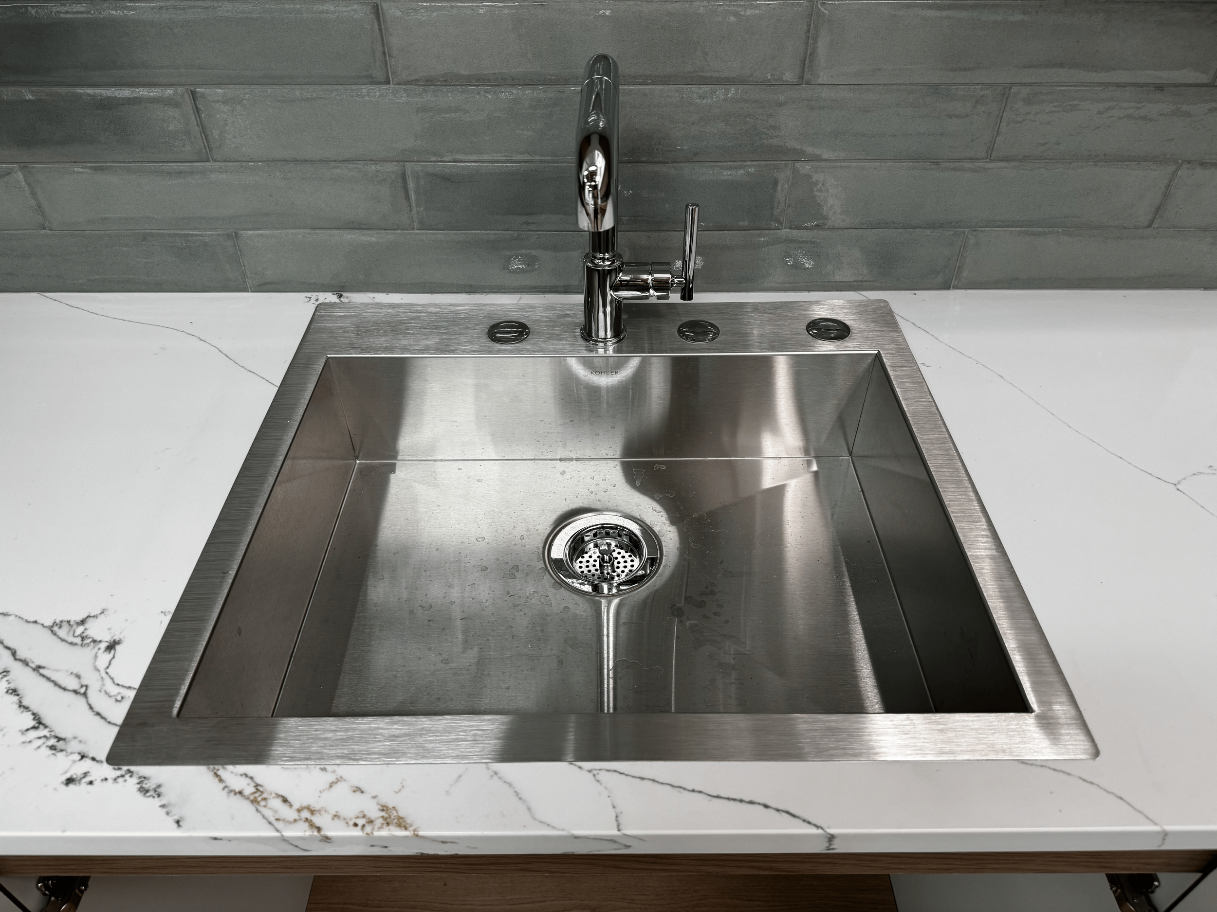 A stainless steel sink with a modern chrome faucet, installed in a white marble countertop with gray veining, against a gray tile backsplash.