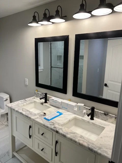Double vanity bathroom with marble countertop, two rectangular sinks, black faucets, two large black-framed mirrors, black wall-mounted light fixtures, and a toilet partially visible on the left.