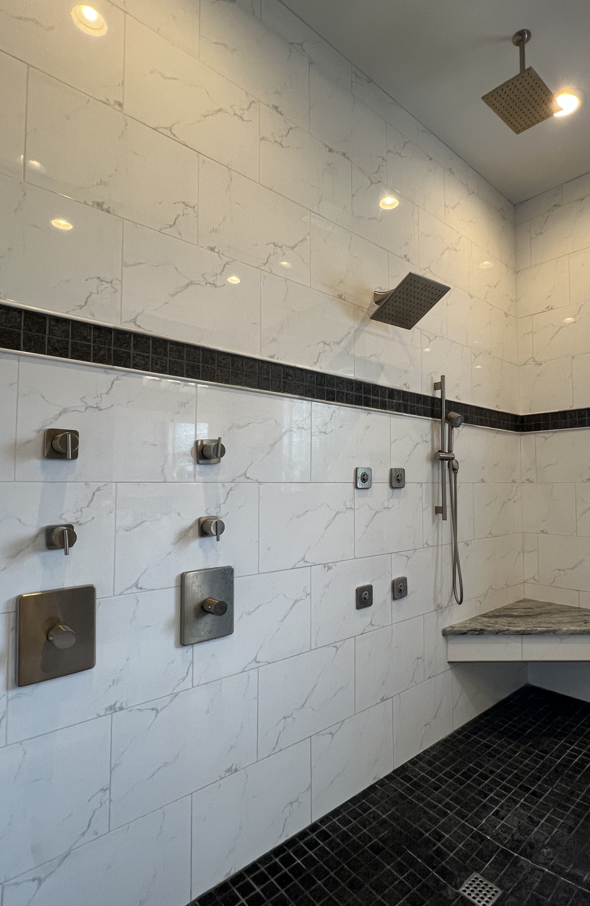 A modern shower with two rainfall showerheads, multiple body jets, and a handheld shower wand, all with brushed metal fixtures on white marble tiles with black accents.
