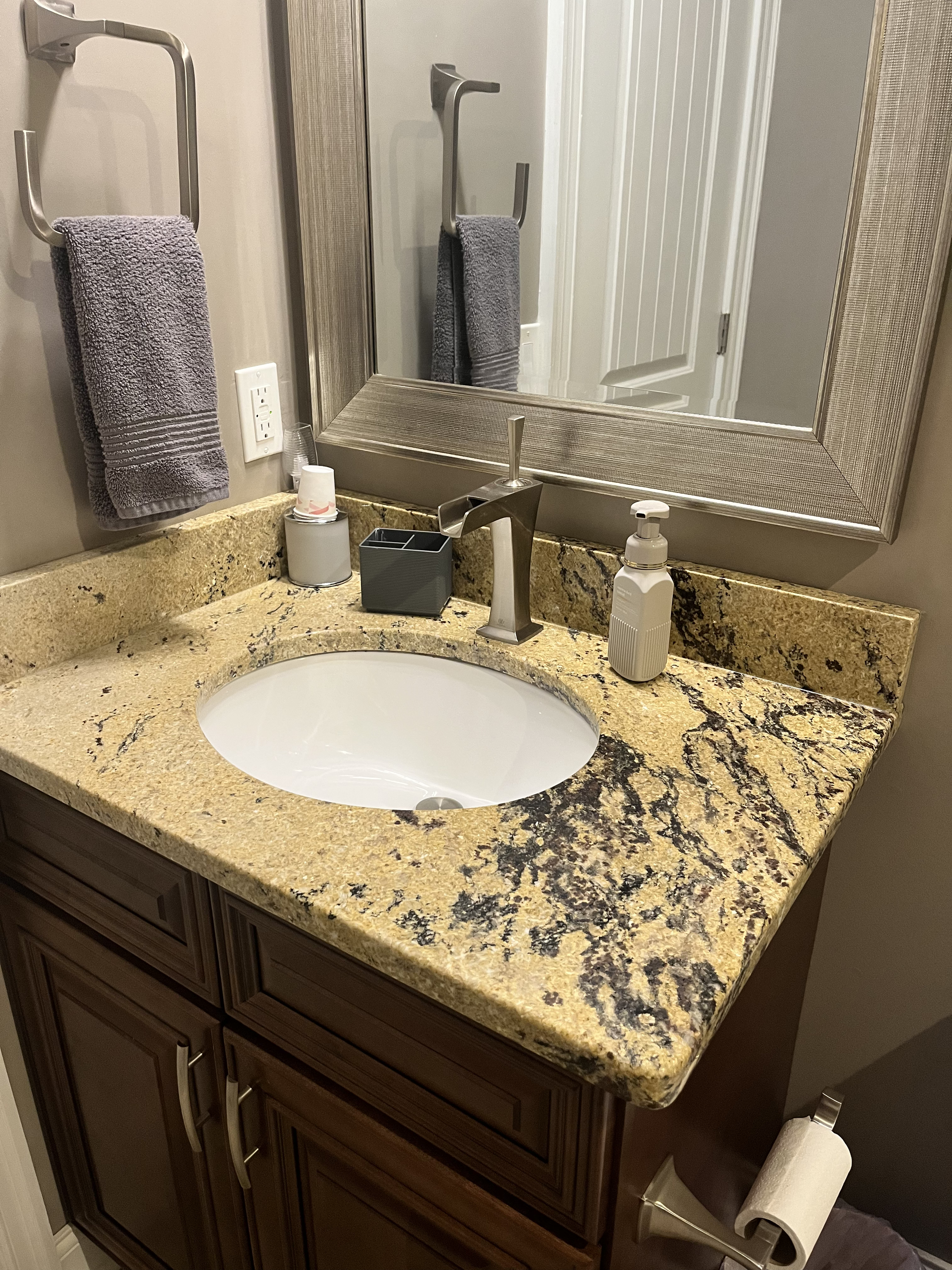 Bathroom vanity with granite countertop, oval sink, faucet, soap dispenser, small container holding cups, wall mirror, towel ring with a lavender towel, and toilet paper holder with toilet paper.