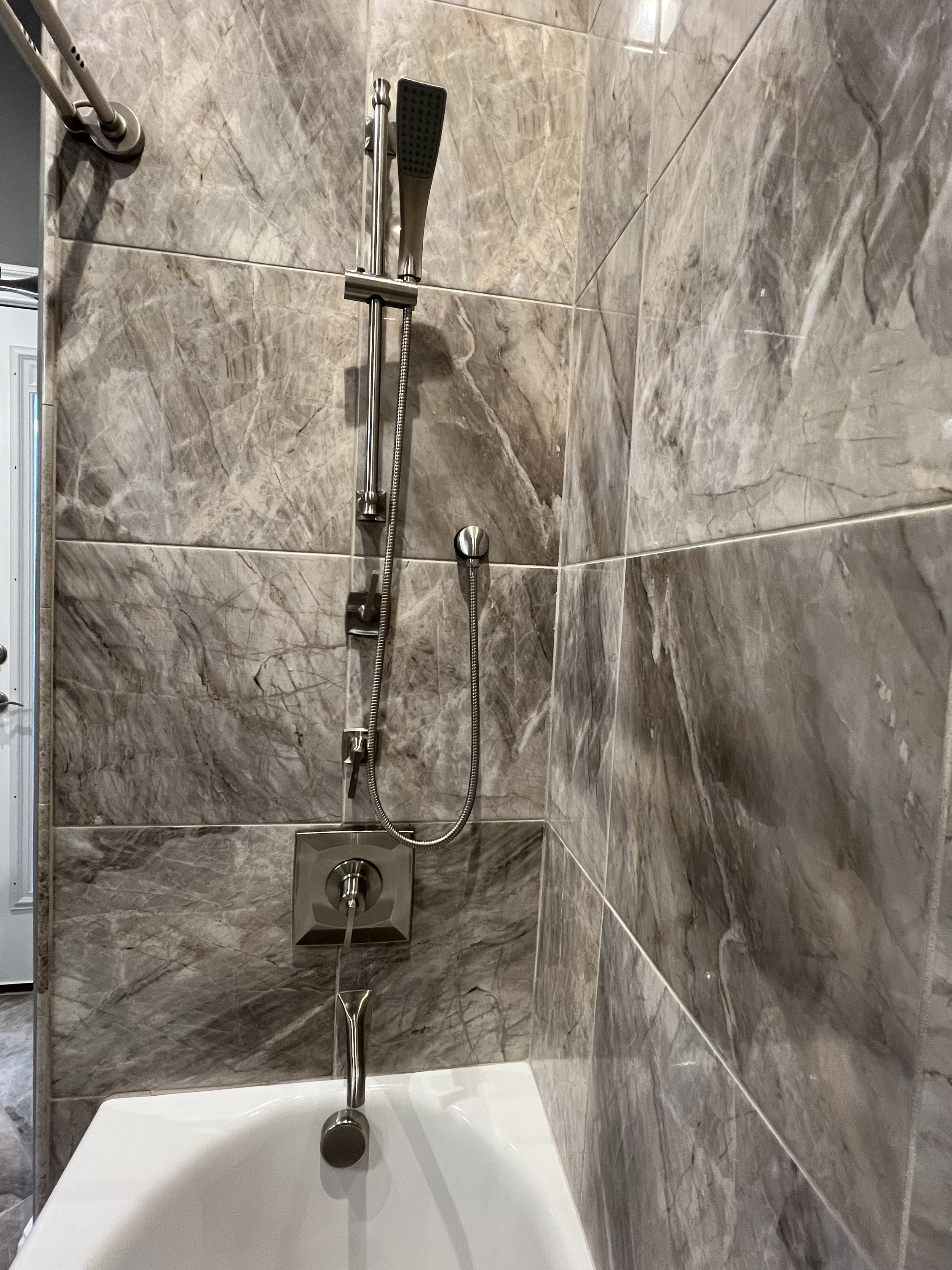 A shower area with gray marble tiles, a handheld showerhead, a wall-mounted square shower control, and a bathtub in the foreground.