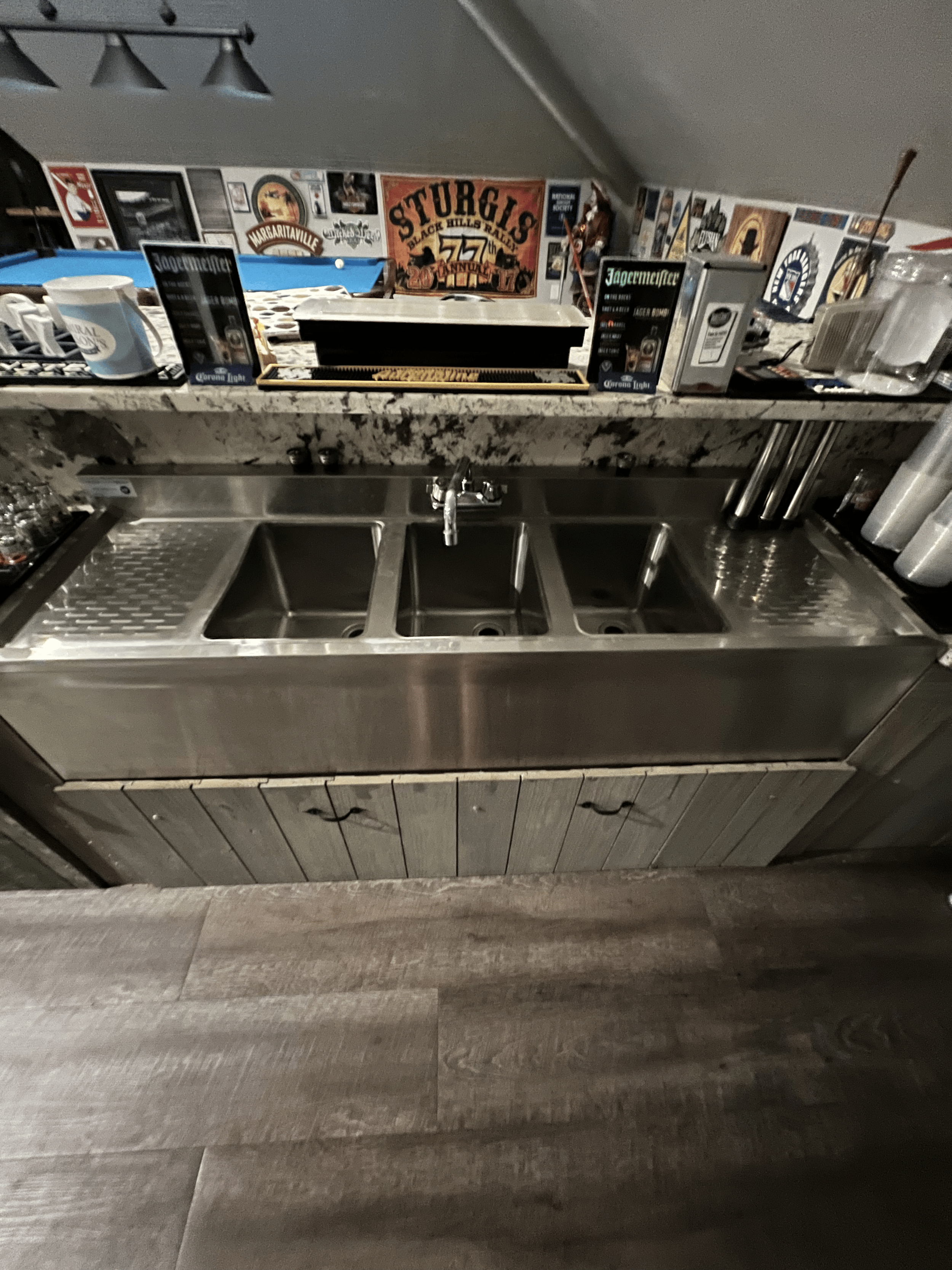 A bar counter with a sink, surrounded by various bar supplies, and a wall decorated with vintage posters and signs.