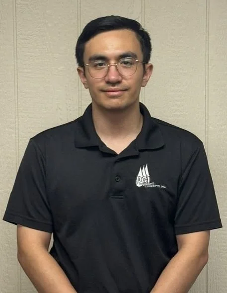 A young man wearing glasses and a black polo shirt with a logo, standing in front of a beige wall.