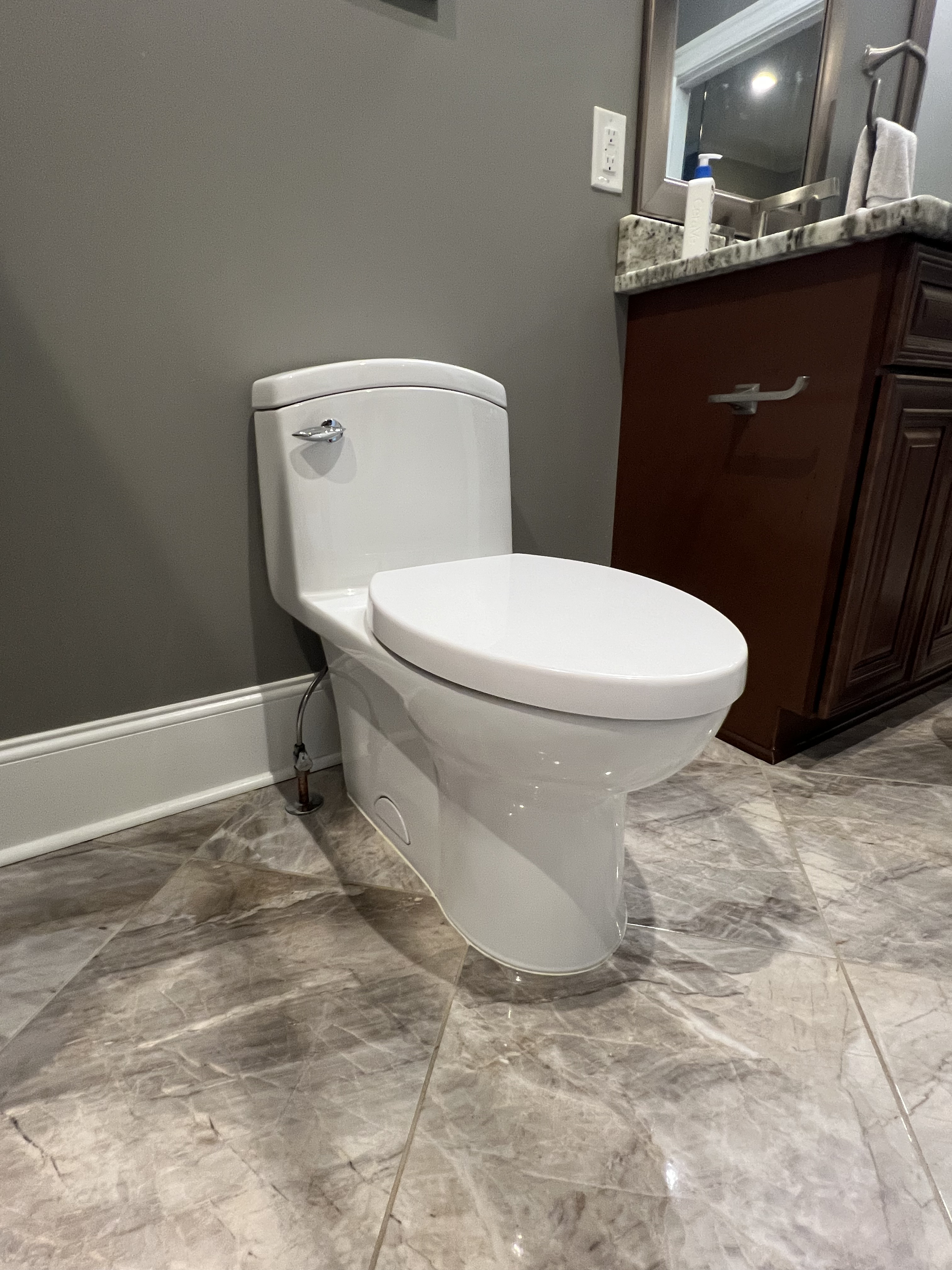 A modern bathroom with a white toilet, dark wooden vanity with a marble countertop, and marble tile flooring. There is a mirror above the sink, a soap dispenser, and a towel hanging on a hook.