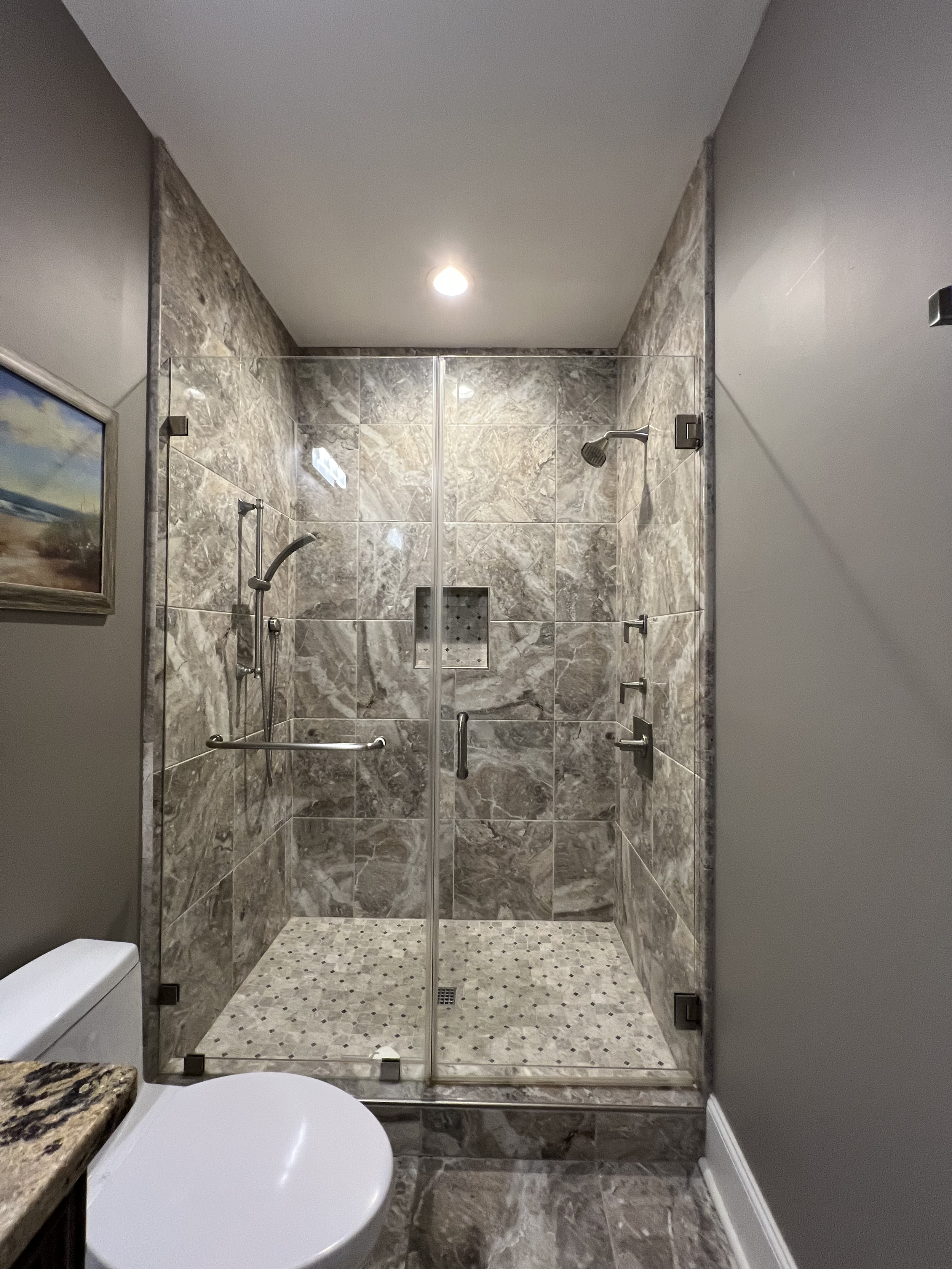 A modern bathroom with a glass shower enclosure featuring marble tiles, a showerhead, and a grab bar. The bathroom has a tiled floor, a toilet in the foreground, and a painting on the wall to the left.