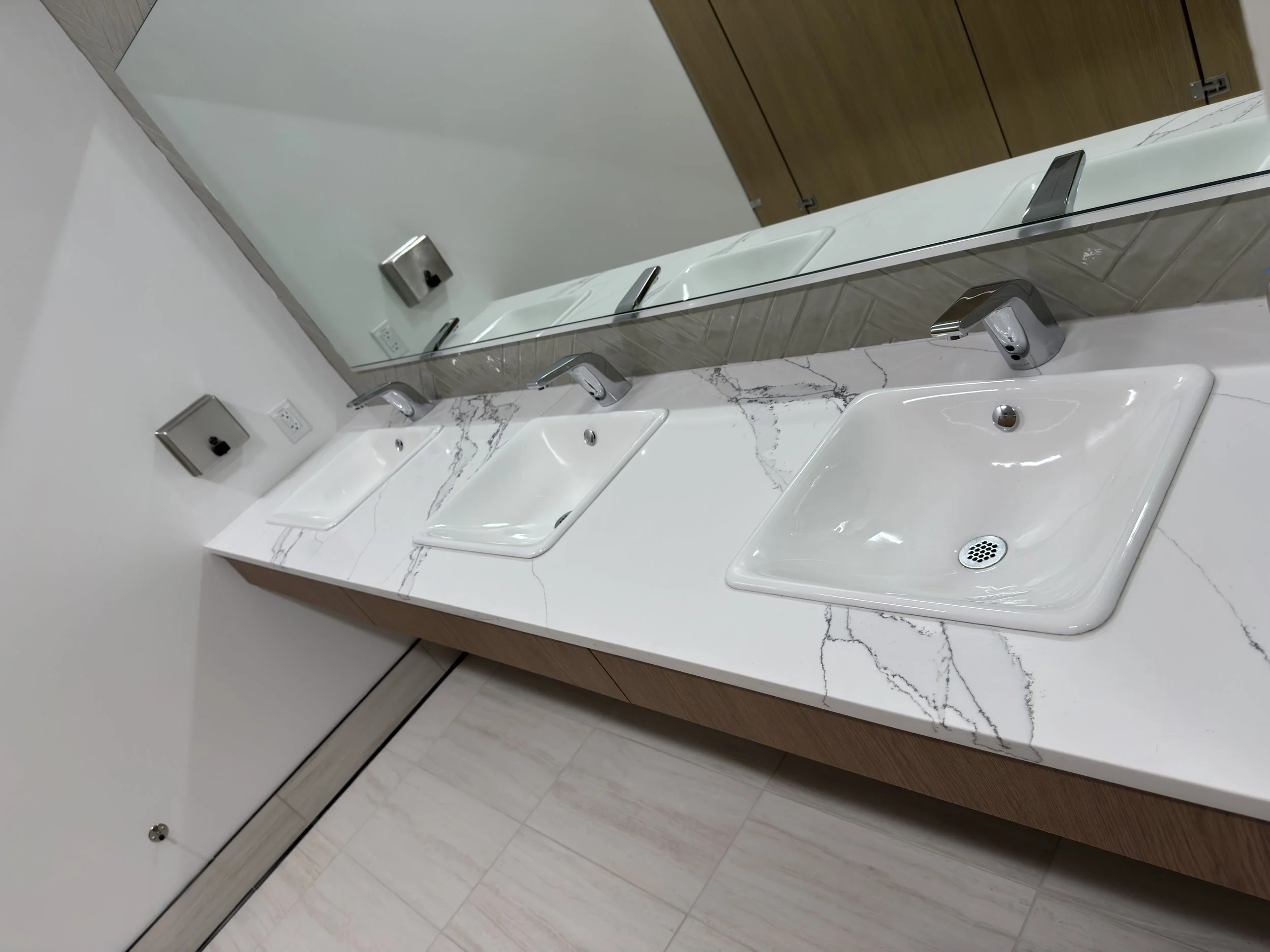 Public restroom sink area with three white sinks, a large mirror, and a wooden cabinet above the sinks.