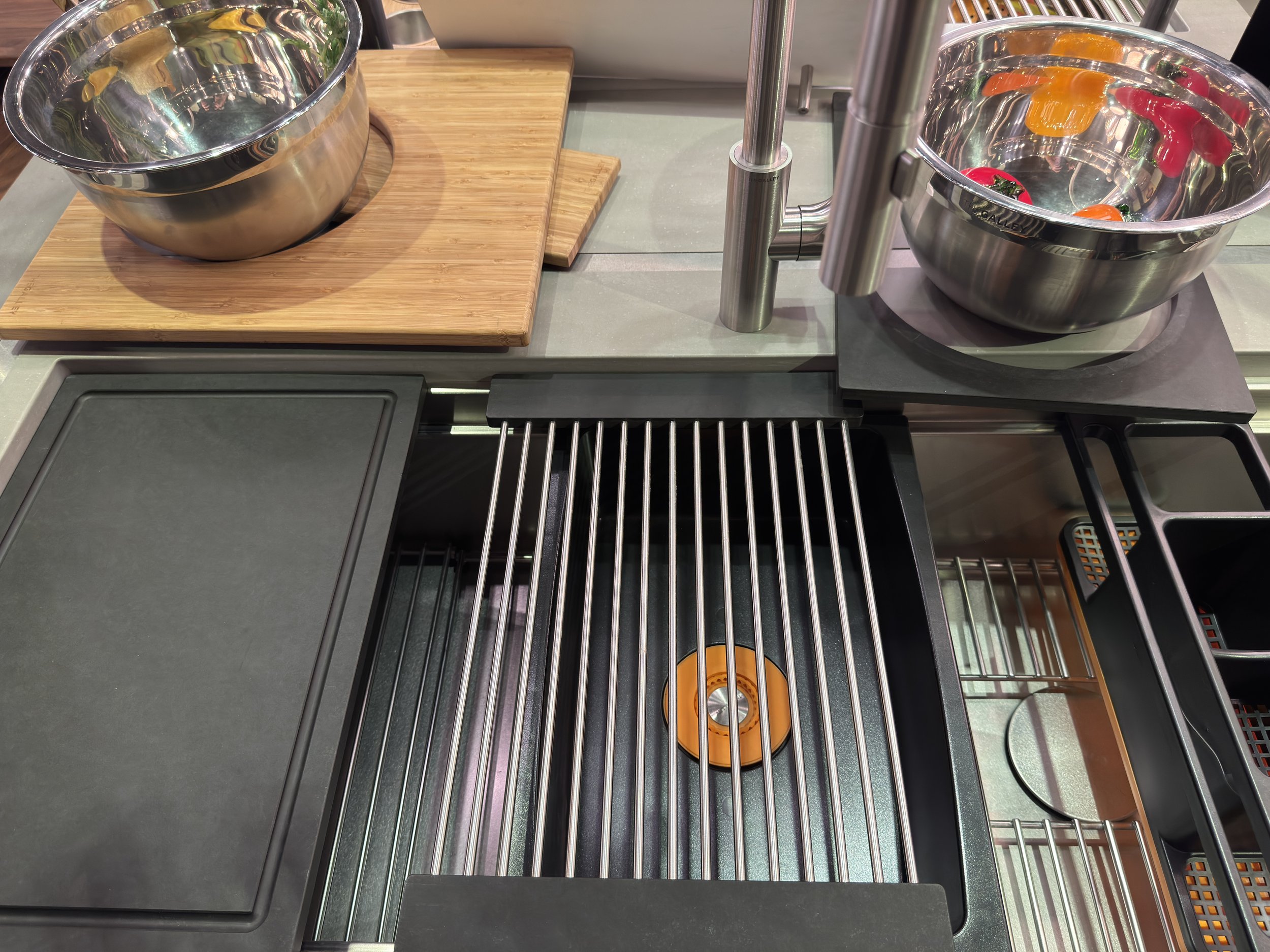 Two stainless steel bowls on a kitchen counter with a wooden cutting board. One bowl contains water and lemon slices, the other bowl contains red, yellow, and orange bell peppers and herbs. A metal faucet is positioned between the bowls.