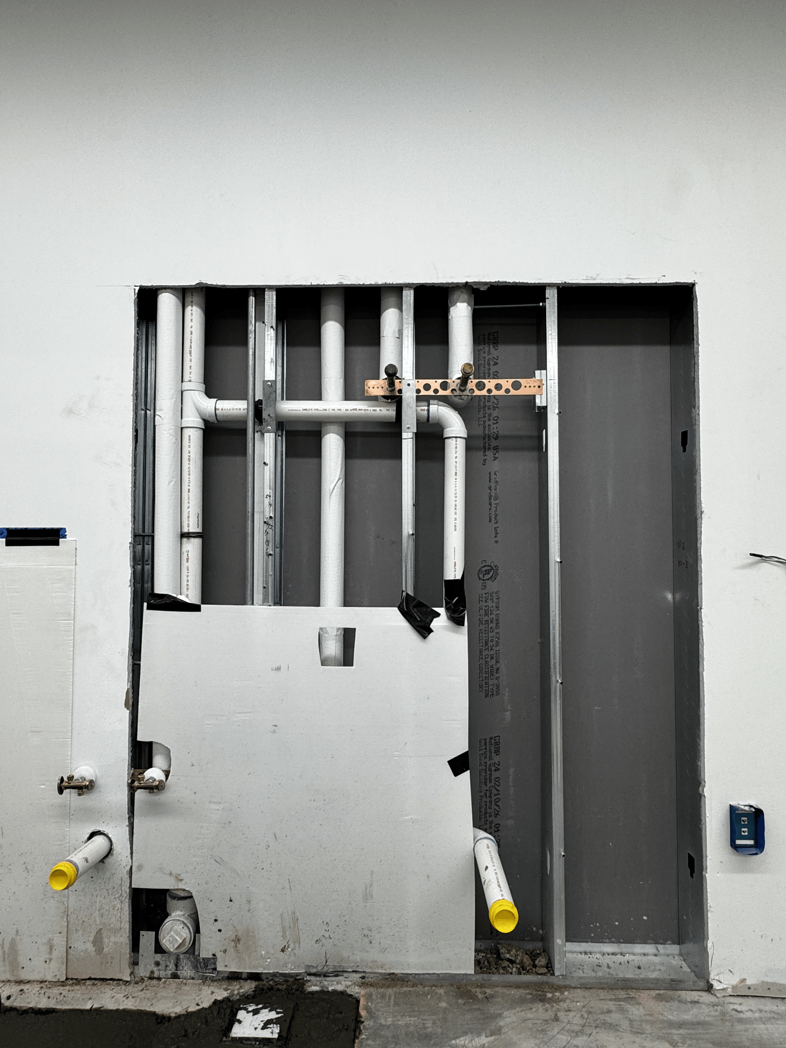Construction site showing an unfinished wall with exposed metal studs and white PVC pipes, some with yellow caps, and a small electrical box on the bottom right.
