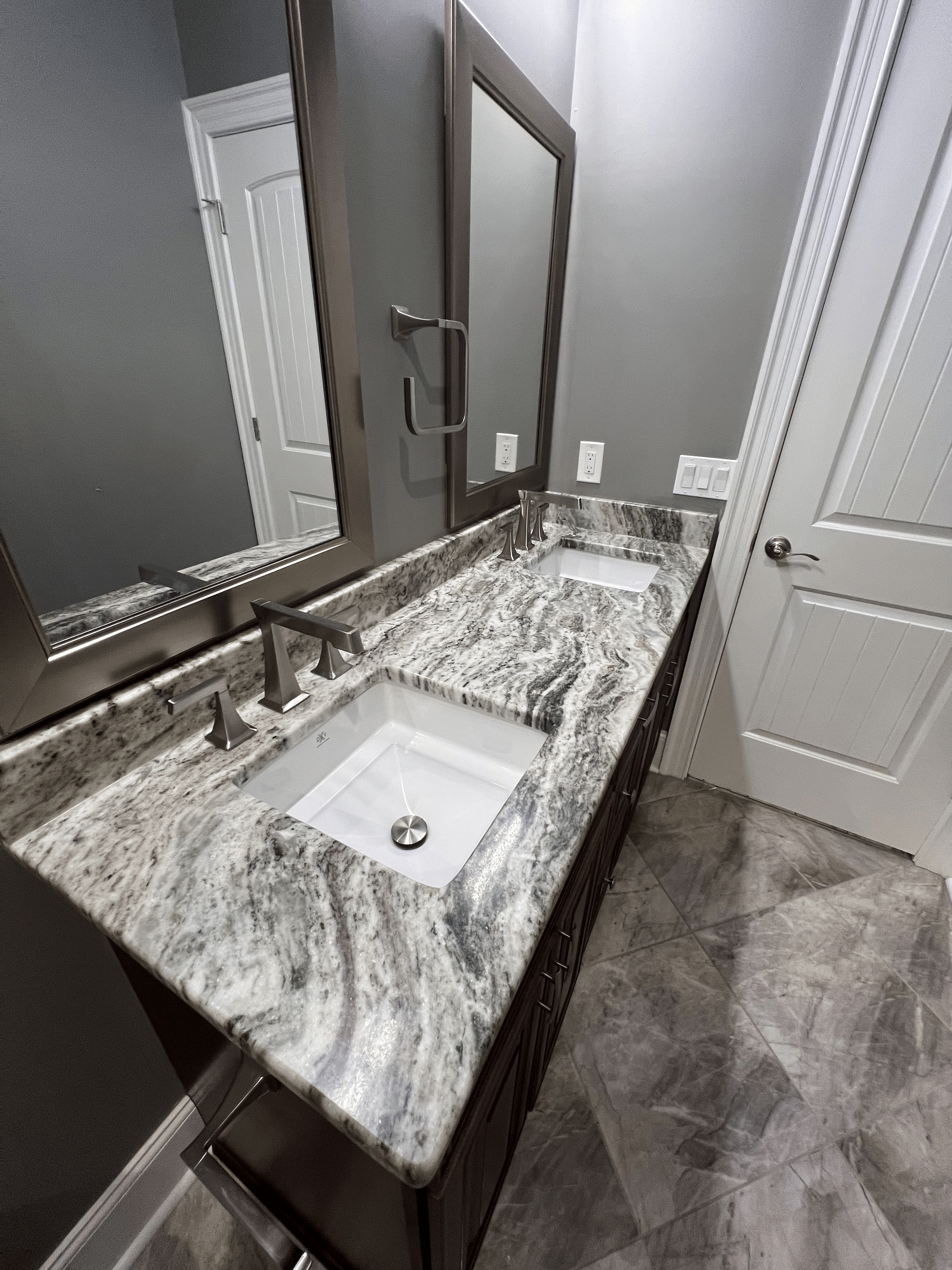 A bathroom double vanity with a granite countertop, two undermount sinks, and modern chrome faucets. Large mirror with three sections is mounted above the countertop, reflecting a door and wall outlets. The bathroom walls are painted gray, and the fl