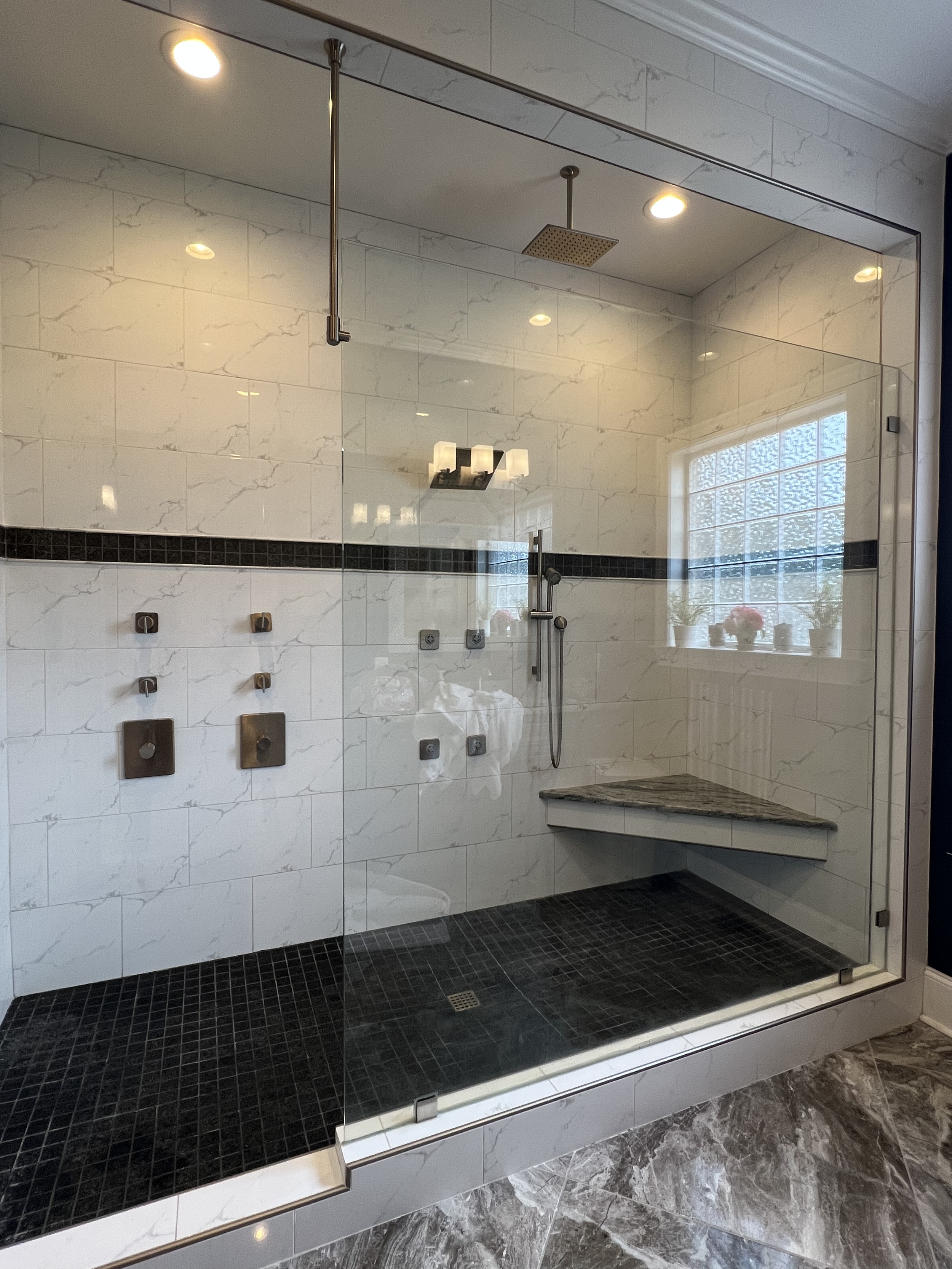 Bathroom shower with glass door, white wall tiles with gray veining, black tiled shower floor, a built-in bench with a marble top, a rain showerhead, and a framed window letting in natural light.