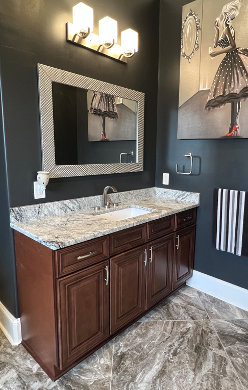 Bathroom vanity area with dark walls, a large framed mirror, a granite countertop with an under-mount sink, dark wood cabinetry, a wall-mounted light fixture, a decorative art piece of a woman in a dress, a towel hanging on a striped towel bar, and p