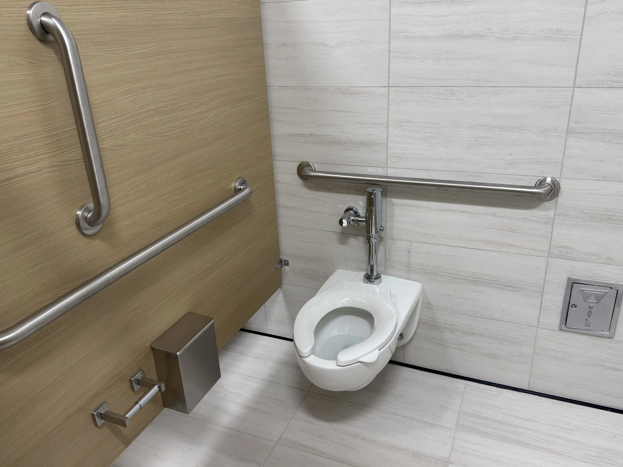 A bathroom with a white toilet, two stainless steel grab bars mounted on the wall, a wooden door, and tiled walls and floor.
