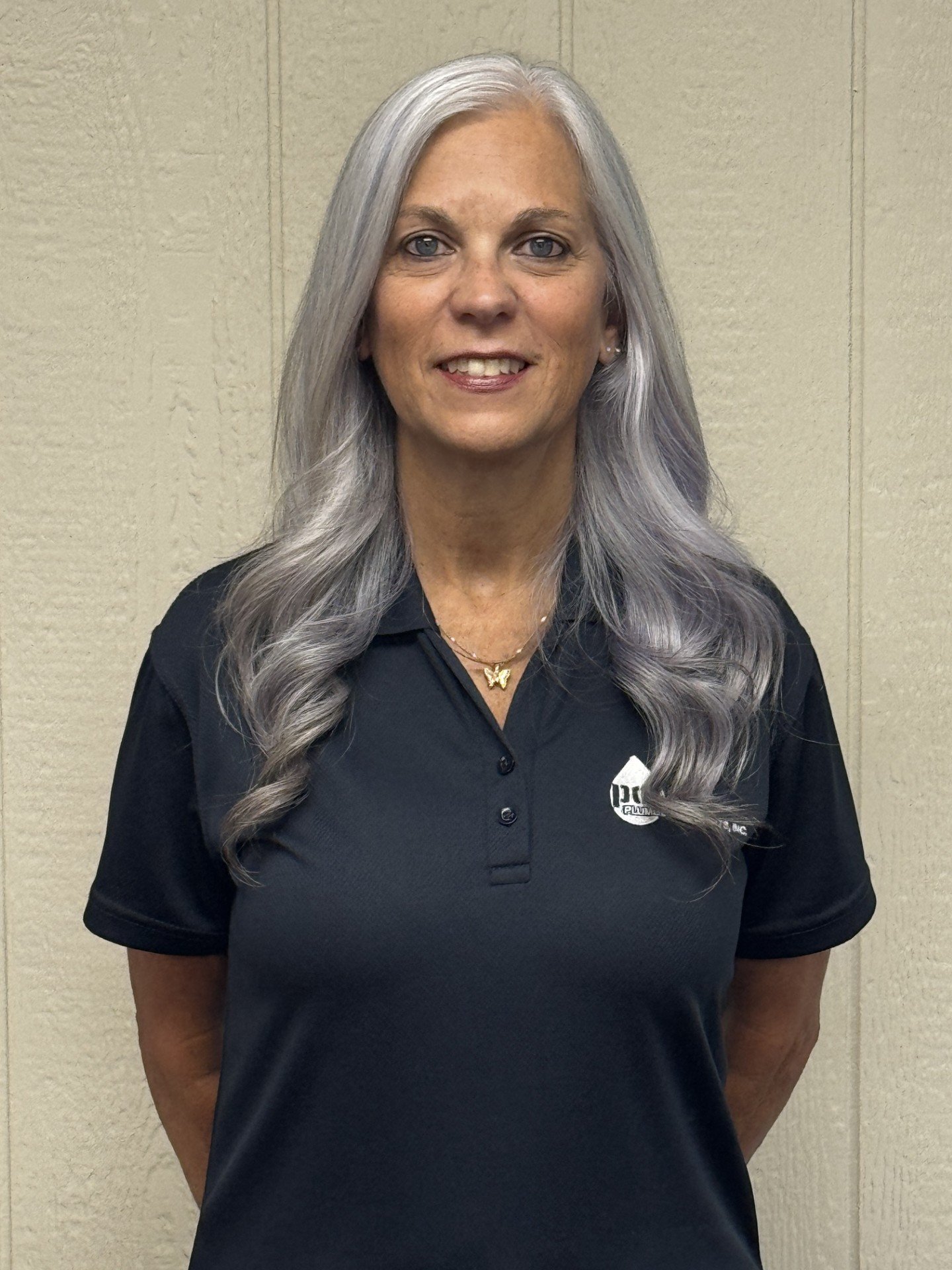A woman with long gray hair, wearing a black polo shirt with a logo on the left side, standing against a beige wall.