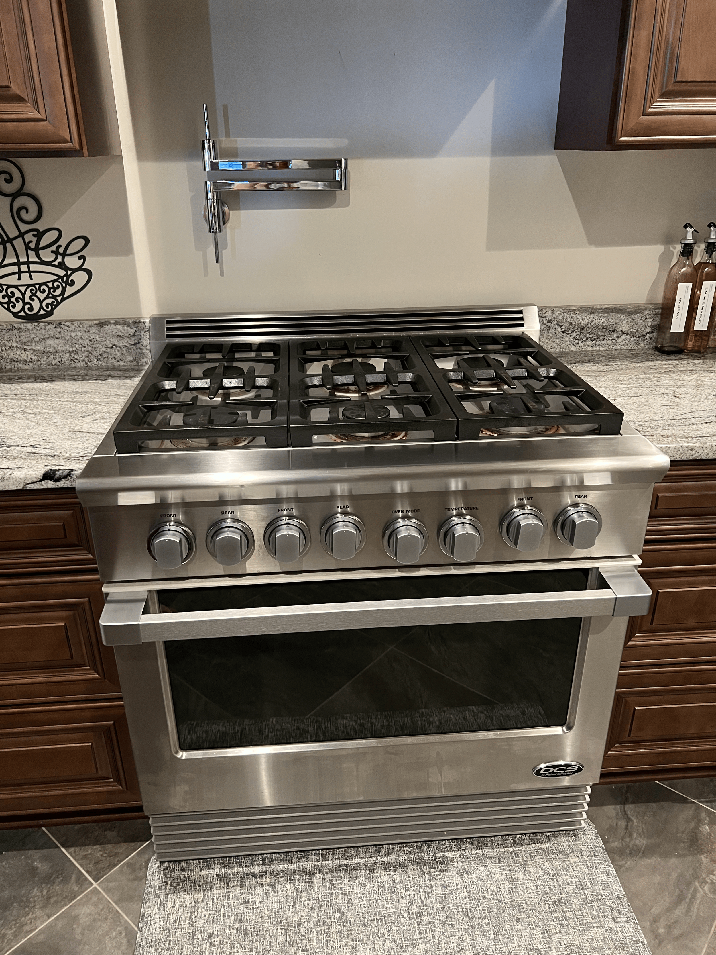 Stainless steel gas stove with six burners, knobs on the front, and oven below in a kitchen with wooden cabinets and granite countertops.