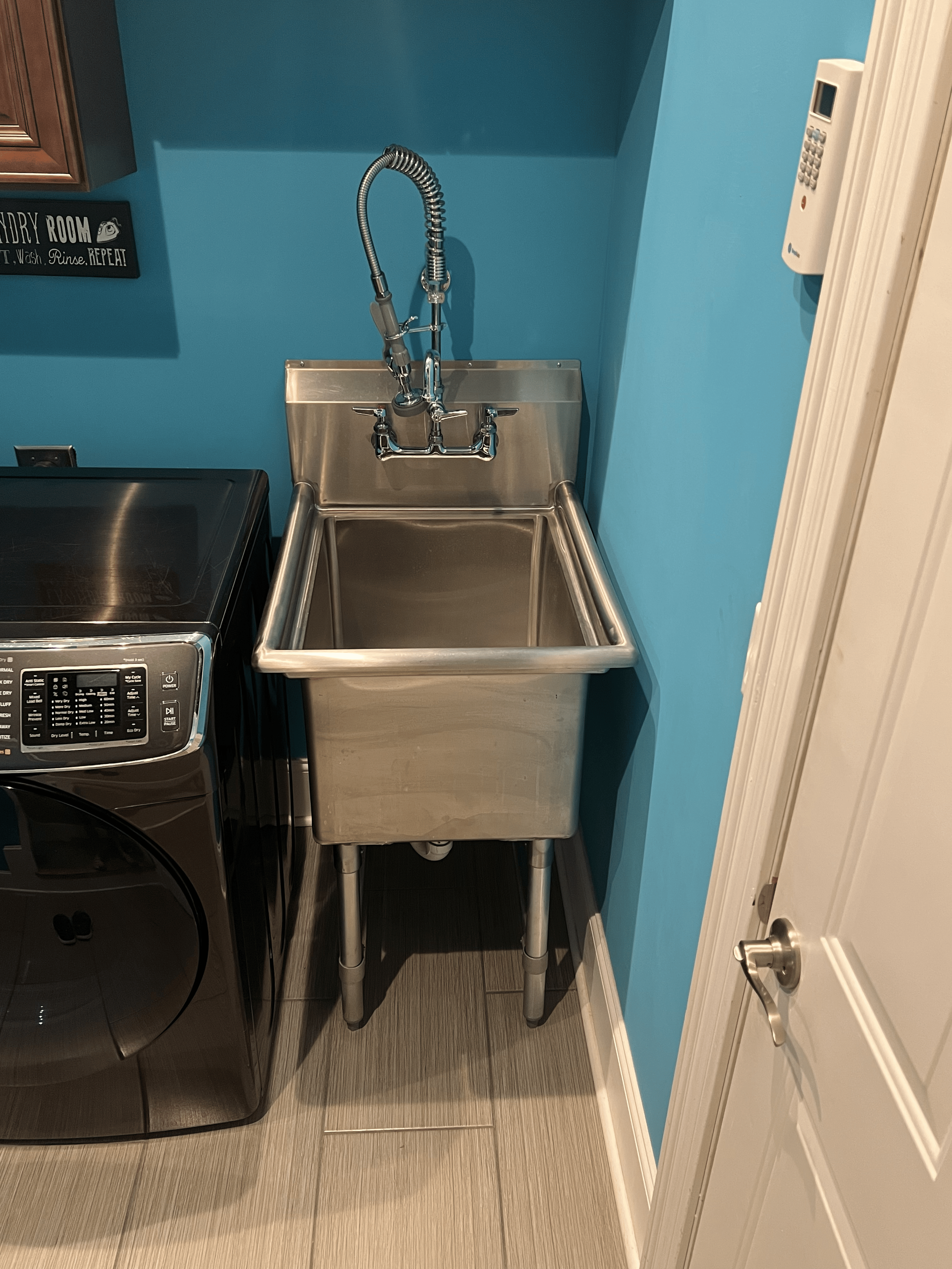 Small stainless steel utility sink against blue wall in laundry room, with a industrial-style faucet and a sign reading 'Laundry Room, T. Wash, Rinse, REPEAT!' to the left, dryer on the left, and white door with silver handle on the right.