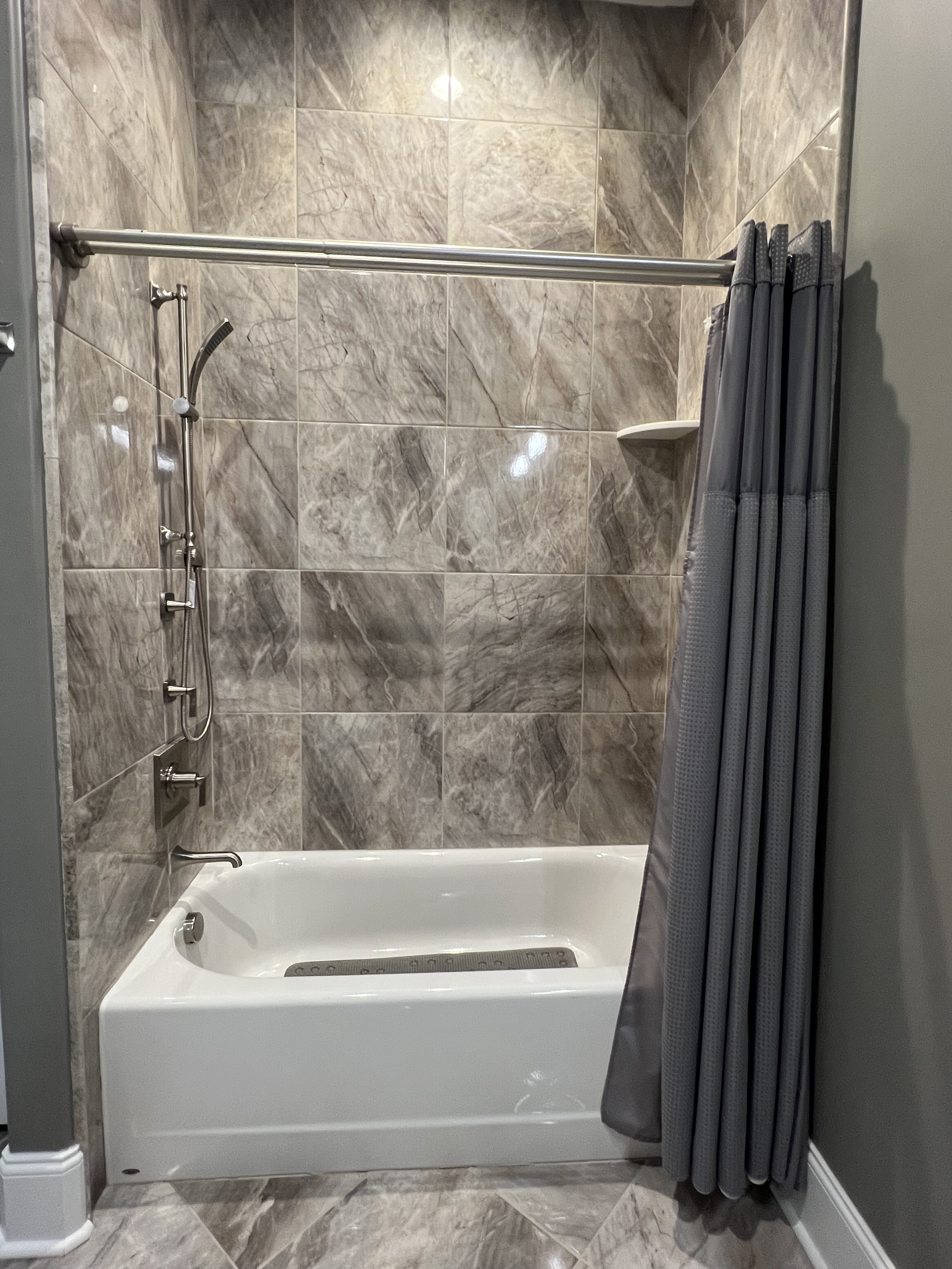 A walk-in shower with gray marble tiled walls, a handheld showerhead, a built-in soap shelf, and a white bathtub with a gray curtain to the side.