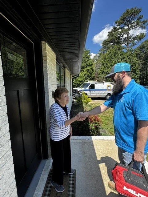 A man in a blue shirt and cap shaking hands with an elderly woman on a front porch, with a police vehicle and trees in the background.