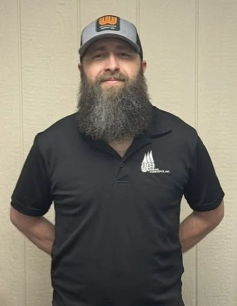 Man with a full beard wearing a baseball cap and black polo shirt standing against a beige paneled wall.