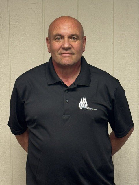 A bald man wearing a black polo shirt with a logo on the left chest standing in front of a beige wall.