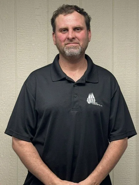 Man with short brown hair and beard wearing a black polo shirt with a logo, standing against a beige wall.