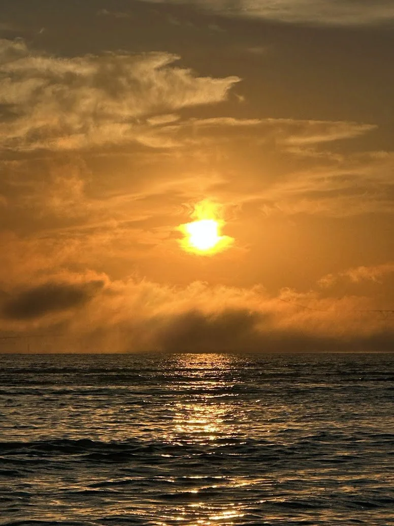 Sunset over the ocean with clouds in the sky, reflections on the water, and the horizon visible.