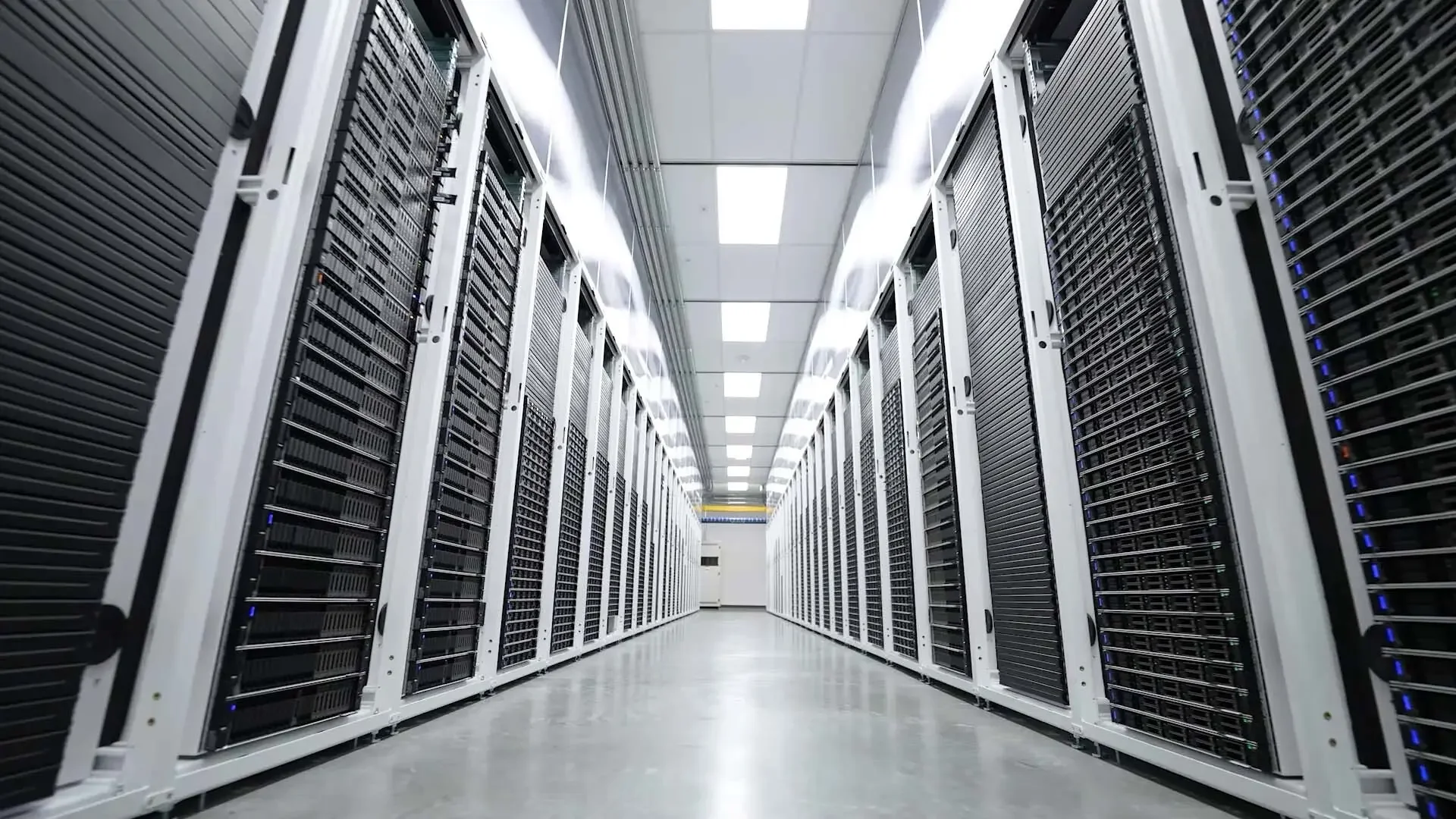 A data center corridor with rows of server racks on both sides and bright ceiling lights.