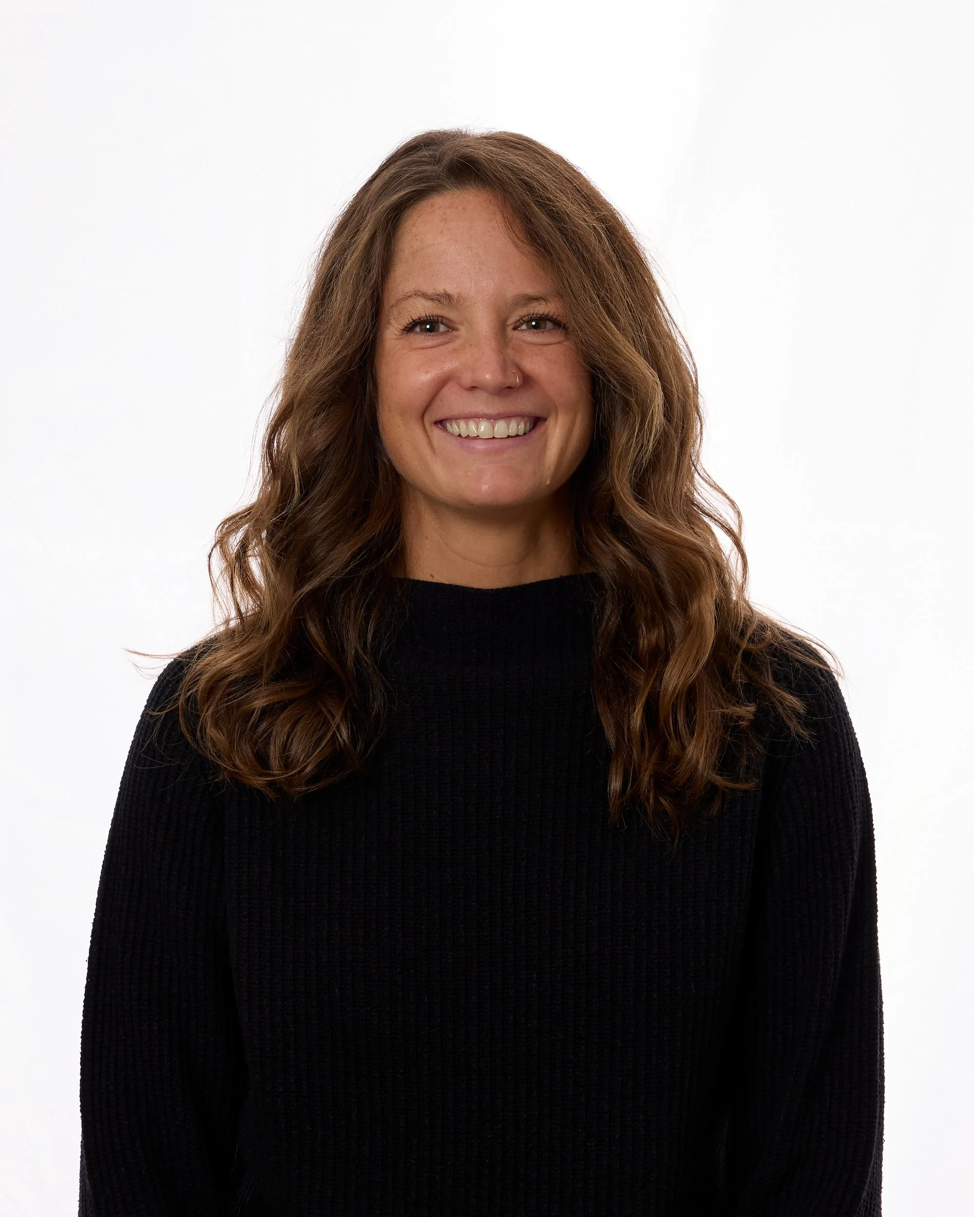 A woman with long, wavy brown hair, smiling, wearing a black sweater, standing against a white background.