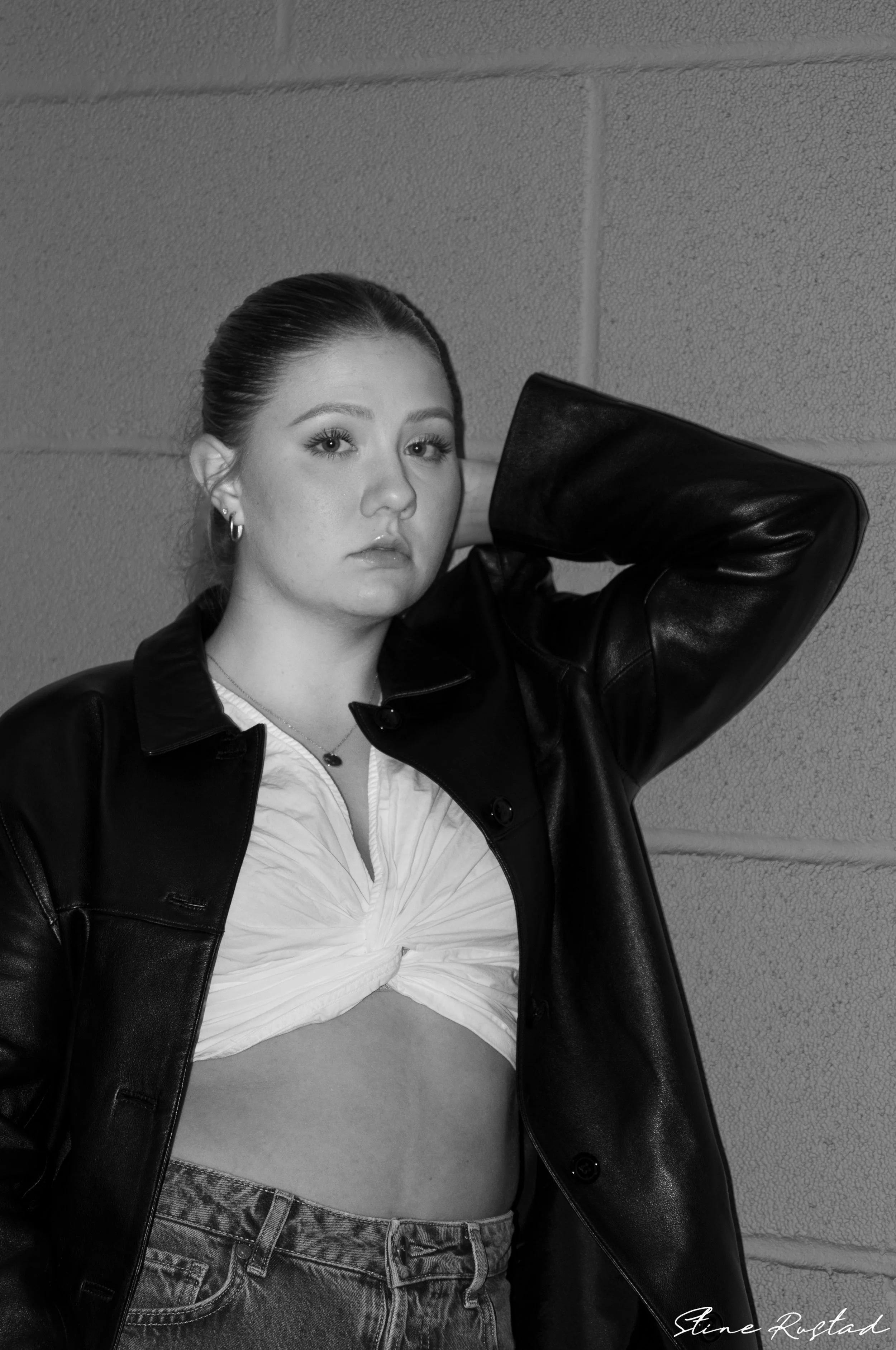 Black and white photo of a young woman with her hair tied back, wearing a leather jacket over a tied-up white shirt and high-waisted jeans, standing against a brick wall.