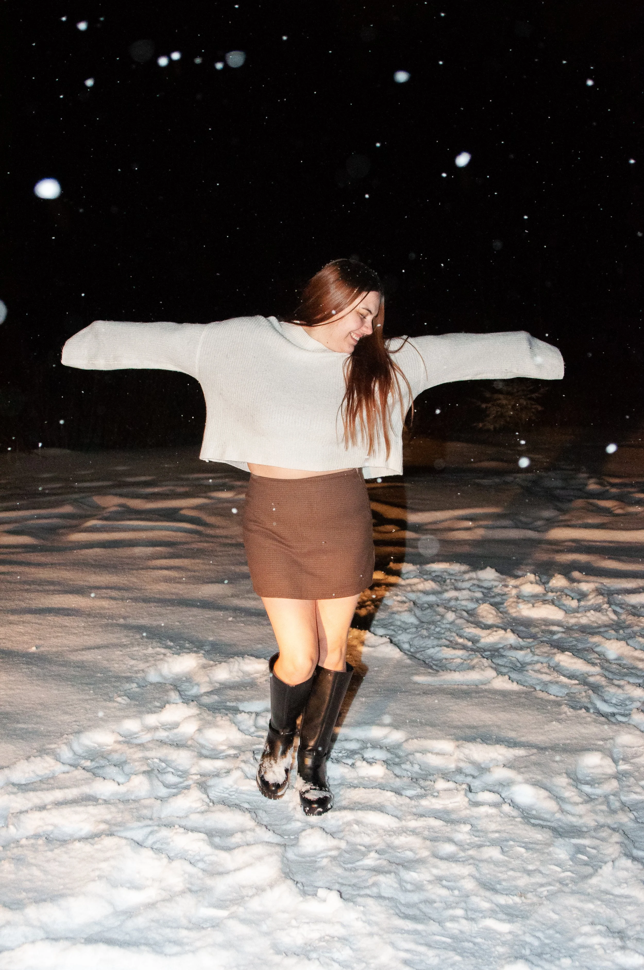 A woman with long hair wearing a white sweater, a brown skirt, and black boots joyfully jumping in the snow at night.