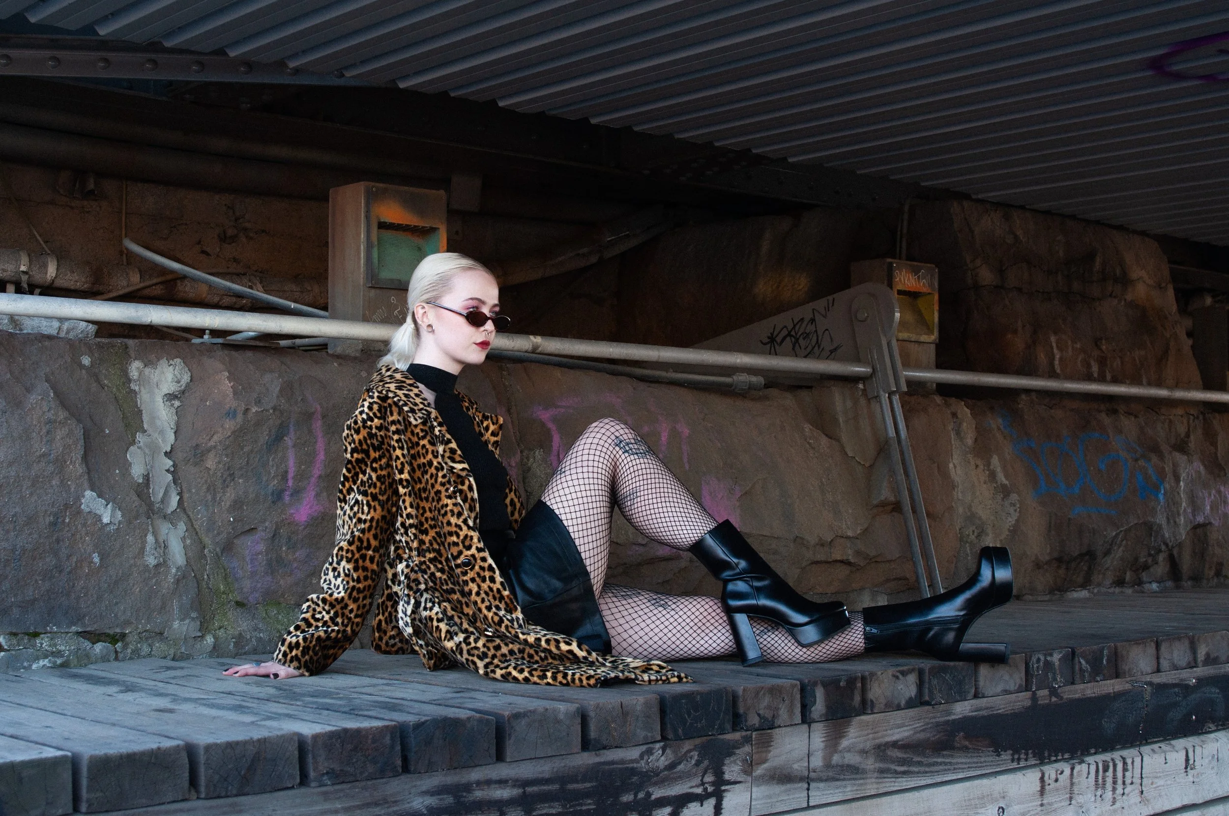 A woman wearing a leopard print coat, black top, fishnet stockings, and black platform boots is sitting on a wooden surface under an overpass. She has platinum blonde hair, small sunglasses, and red lipstick, with graffiti and rocks in the background