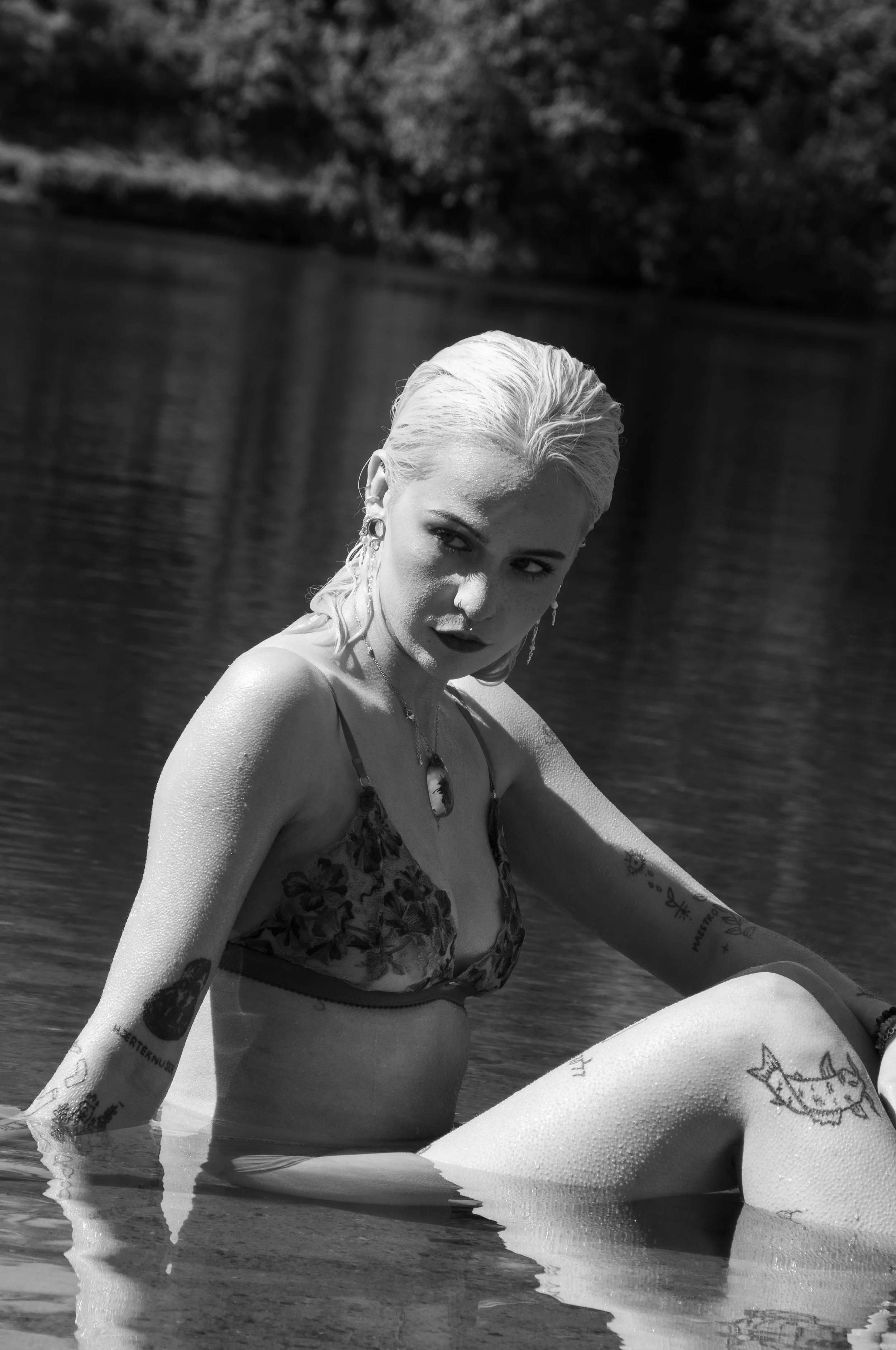 A black-and-white photo of a woman with wet blonde hair sitting in a body of water, wearing a floral bikini top and various tattoos.