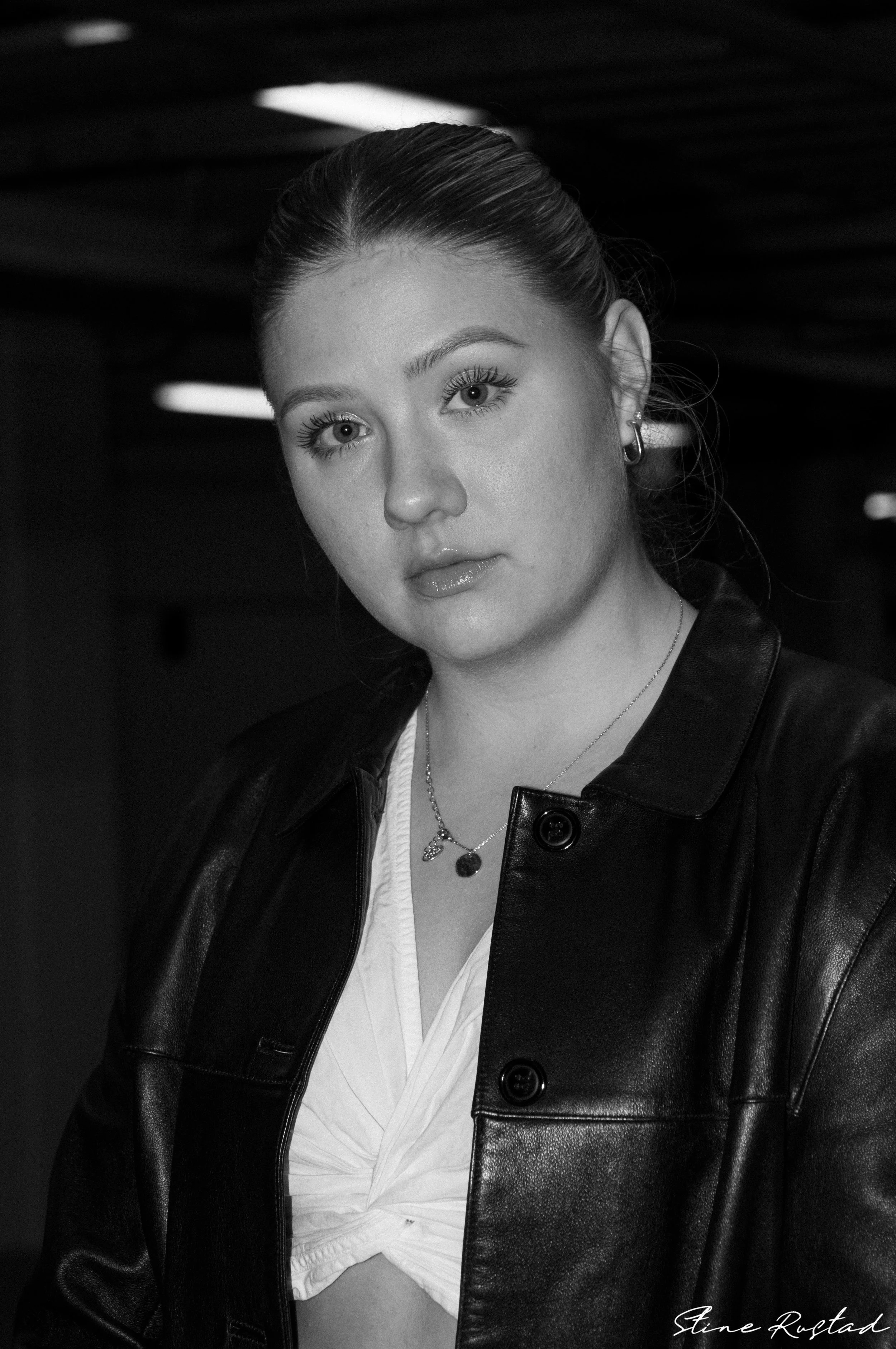 A portrait of a young woman with light skin, wearing a leather jacket and a white top, with her hair pulled back. She is looking directly at the camera with a neutral expression.