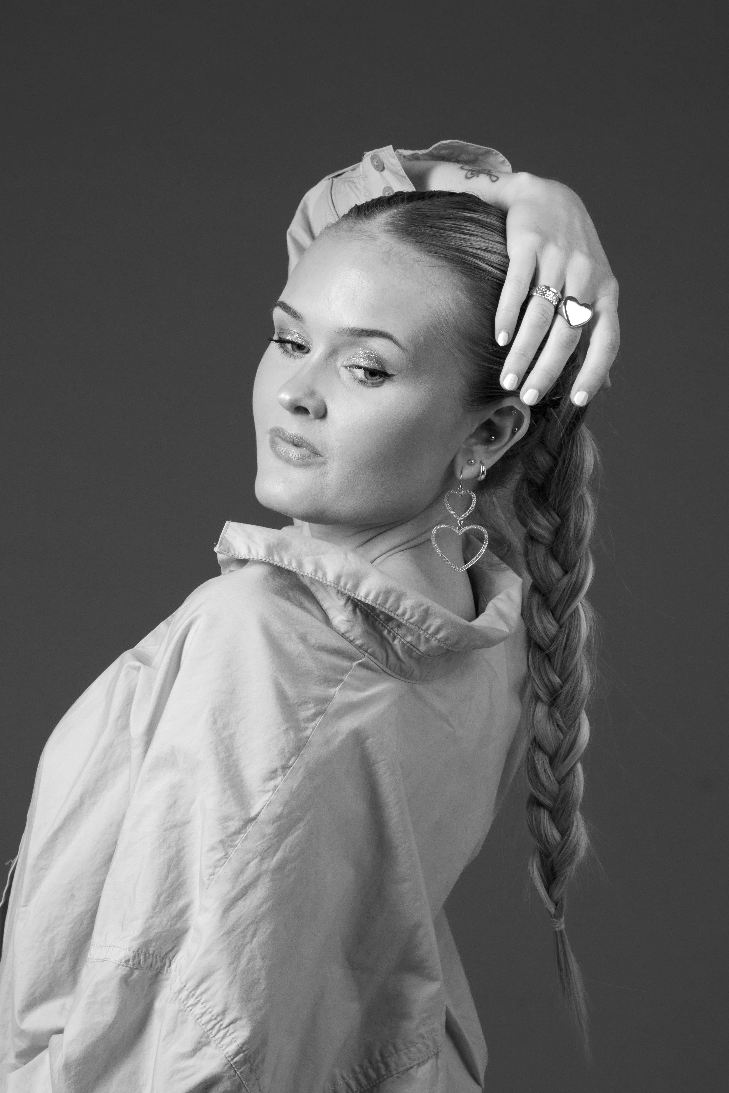 Black and white photo of a woman with a braided hairstyle, wearing heart-shaped earrings, a ring, and a loose jacket, posing with her hand on her head against a plain background.