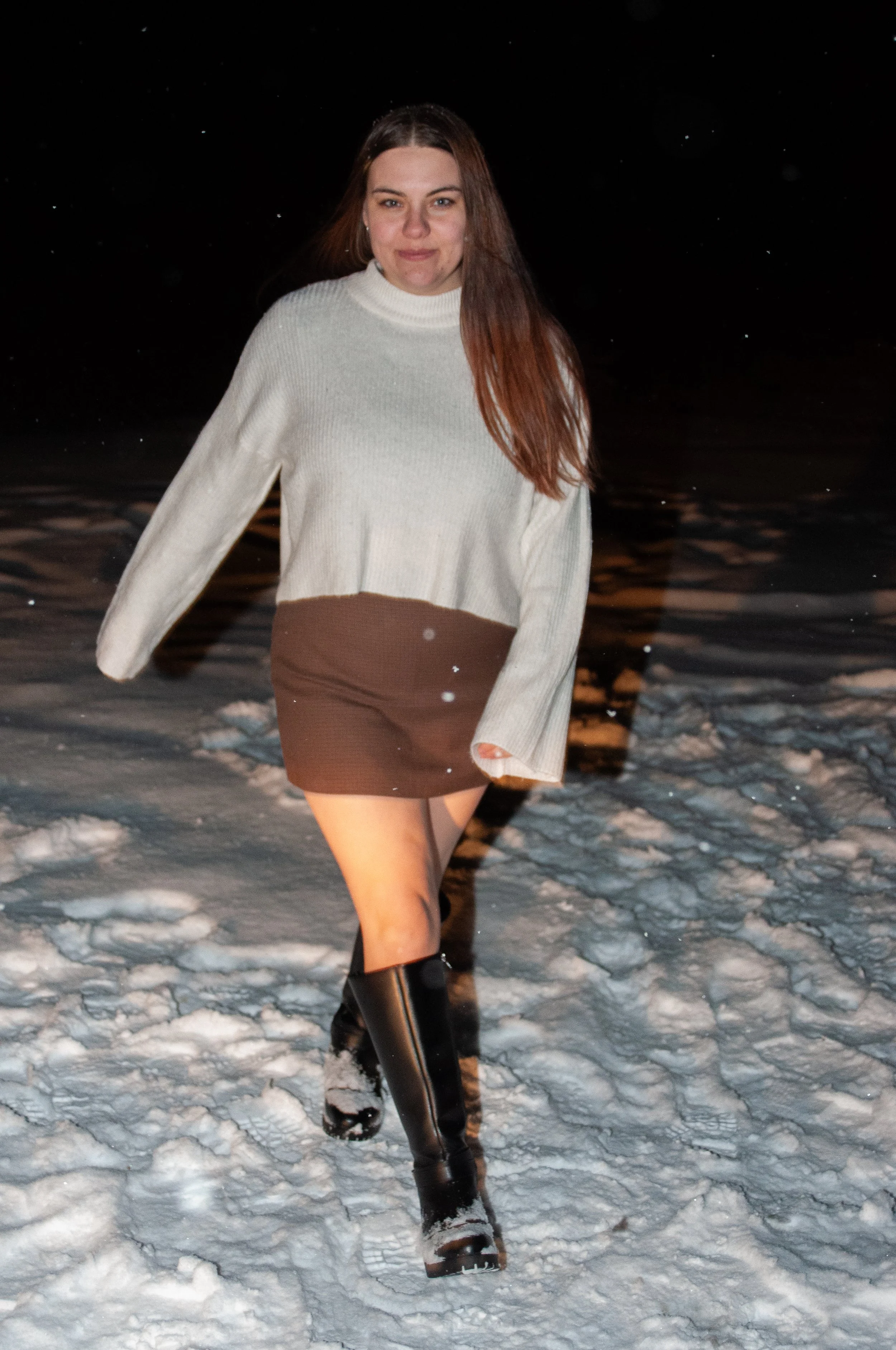 A woman walking outside at night on snowy ground, wearing a white sweater, brown skirt, and black thigh-high boots.