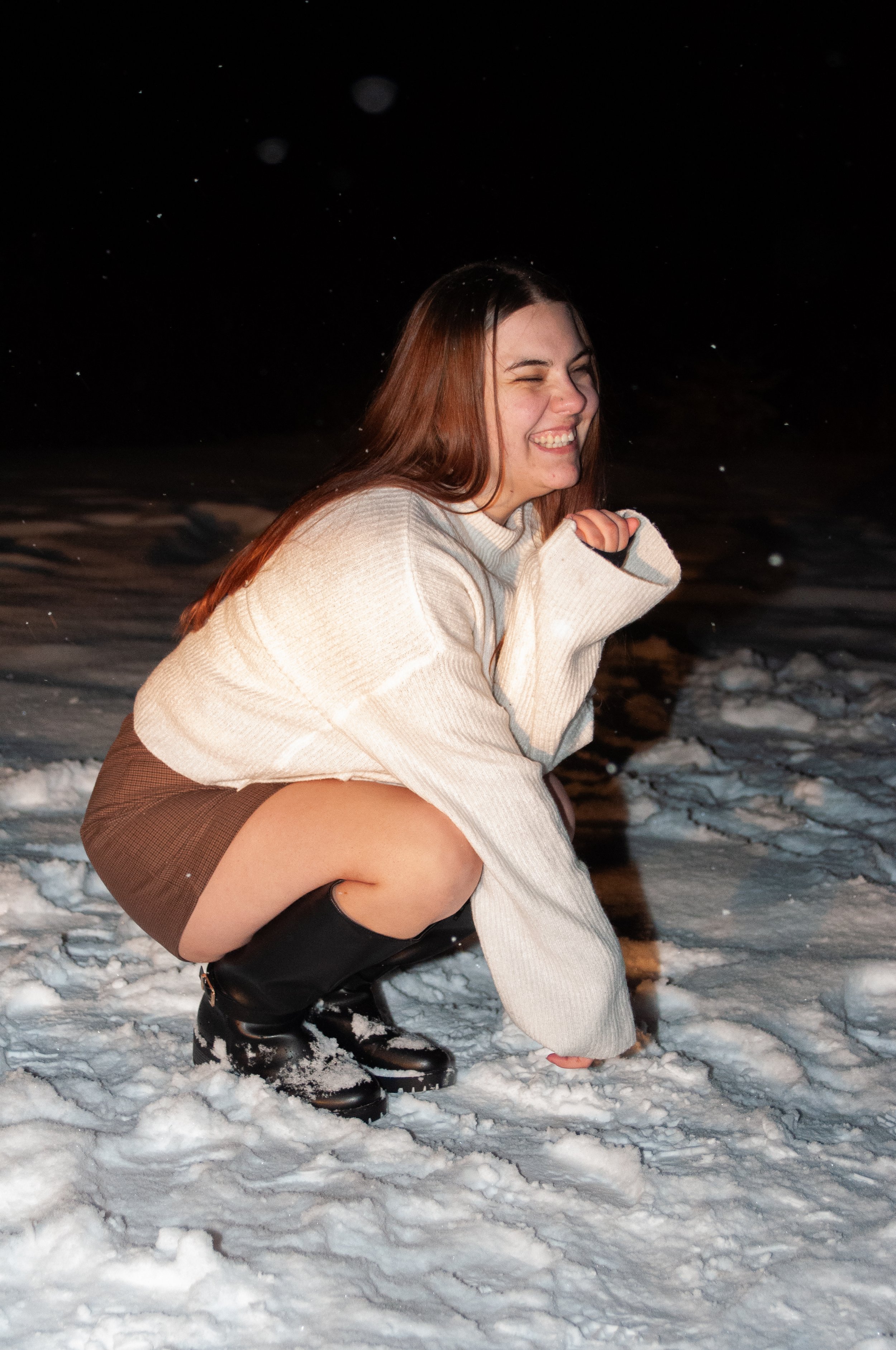 A young woman with brown hair squatting in the snow at night, smiling and laughing, wearing a cream sweater, brown skirt, and black boots.