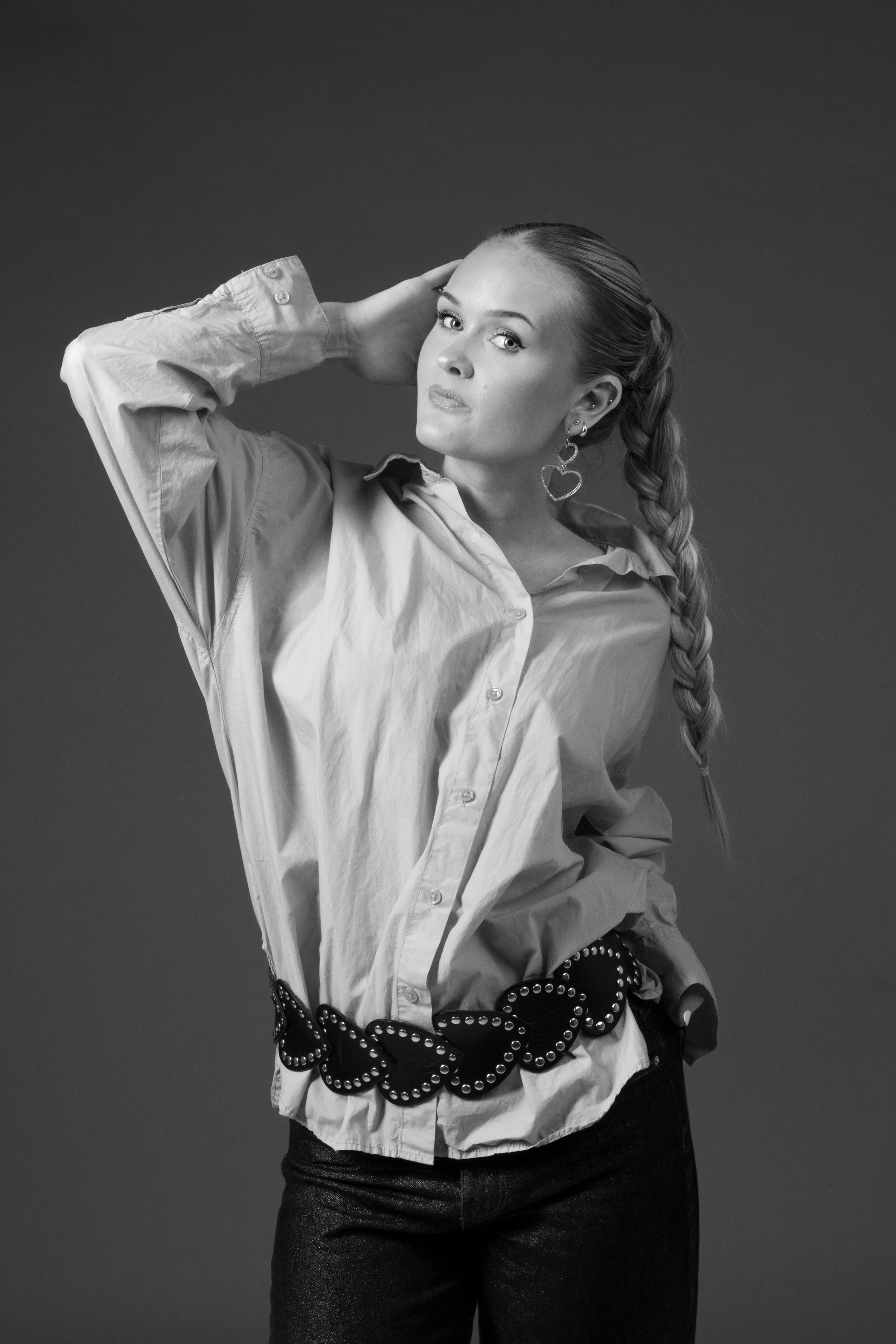 A woman with long braided hair wearing a loose button-up shirt, patterned belt, and earrings, posing with one hand behind her head against a plain background.