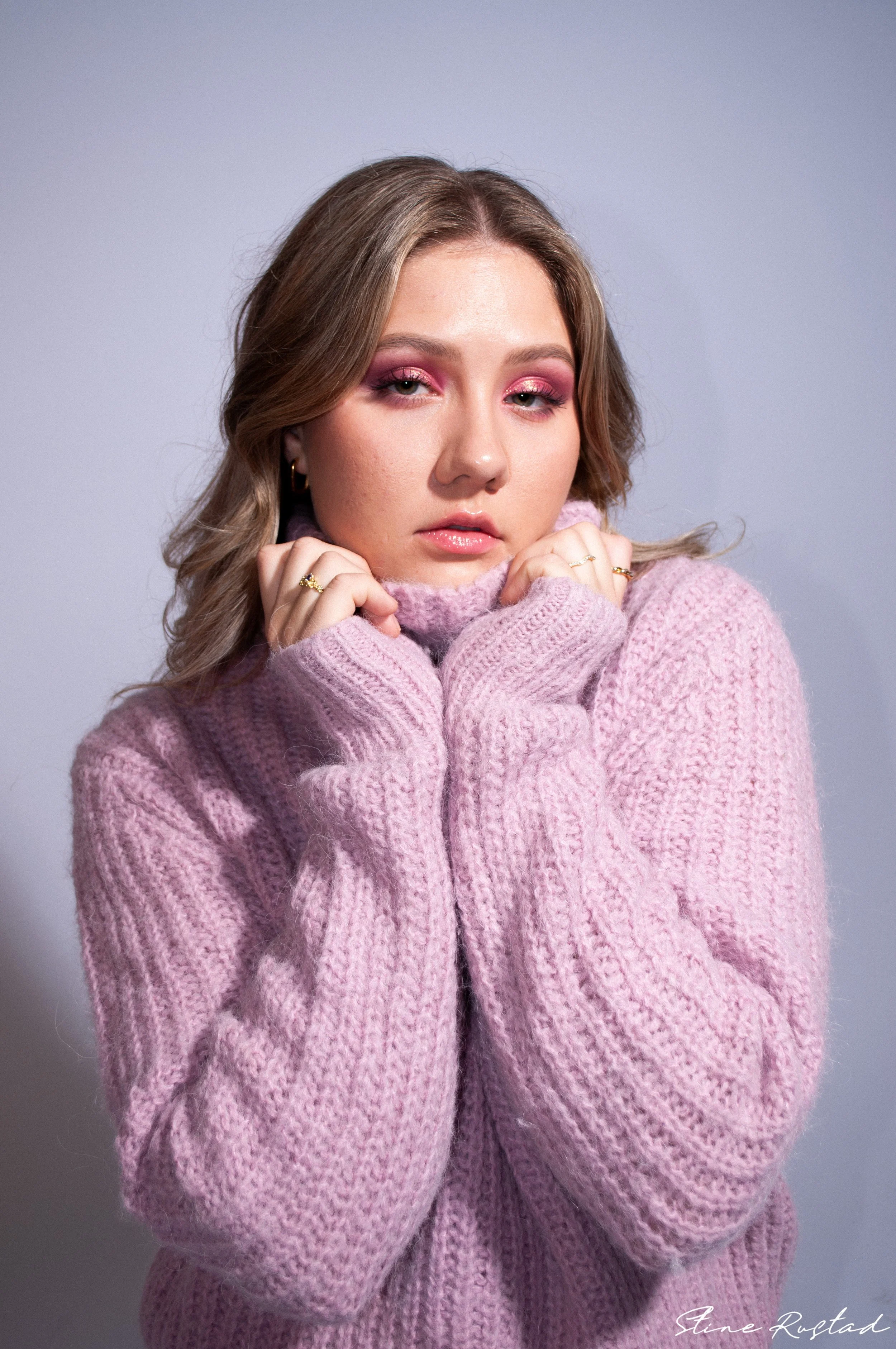 Young woman with light brown hair and pink eyeshadow wearing a pink chunky knit sweater, holding the collar close to her face, looking at the camera.