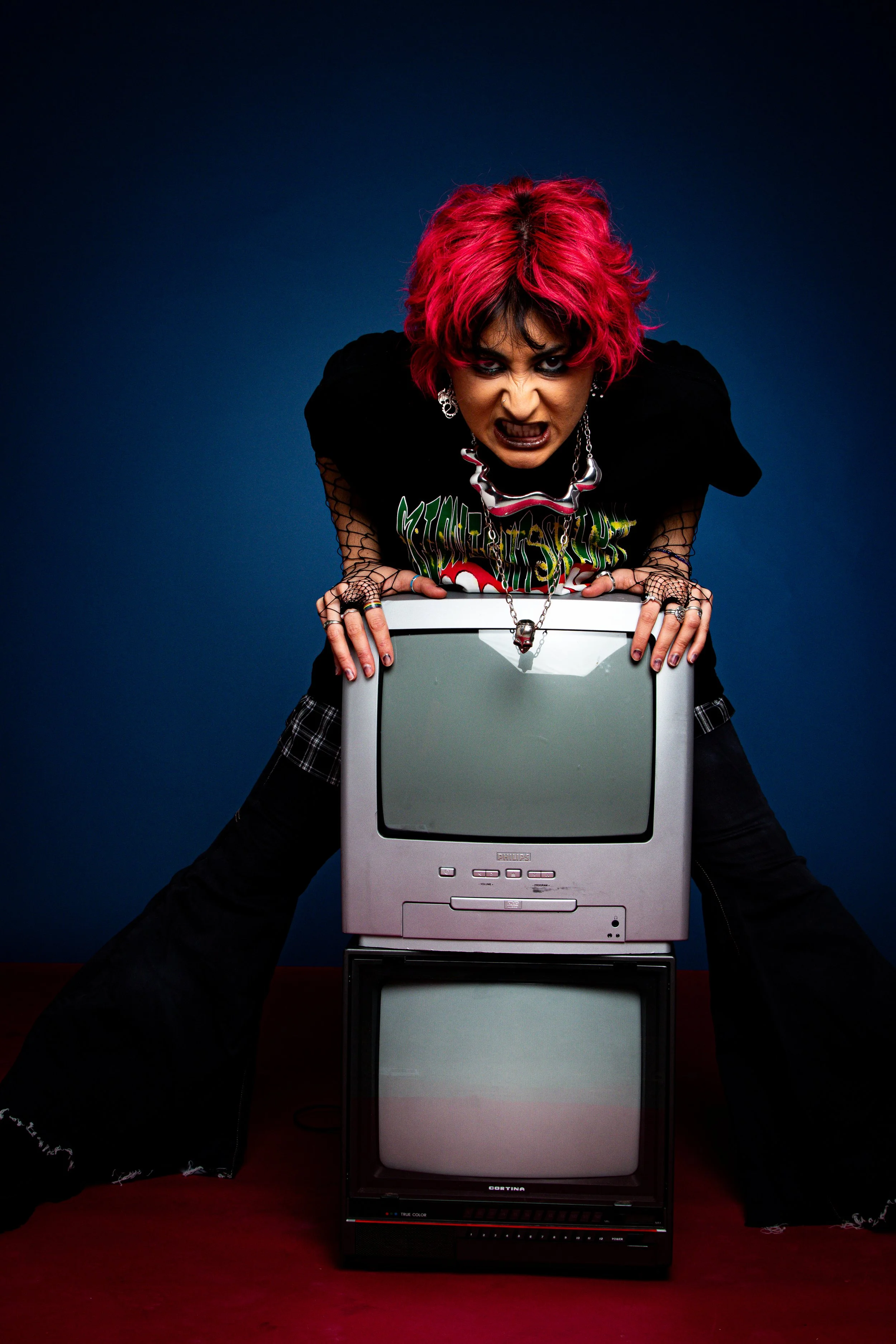 A person with bright pink hair, dark makeup, and gothic clothing leans over two stacked vintage televisions, making an angry or intense facial expression.