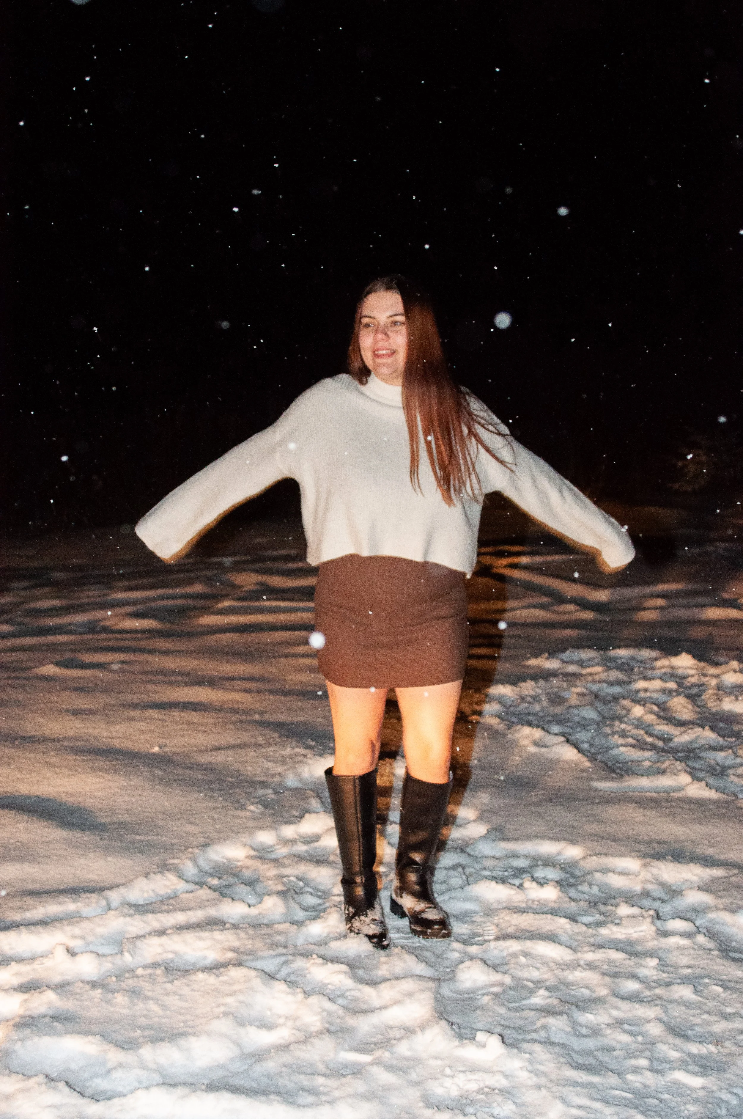 Woman standing in snow at night, wearing a cream sweater, brown mini skirt, and tall black boots, with snow falling around her.